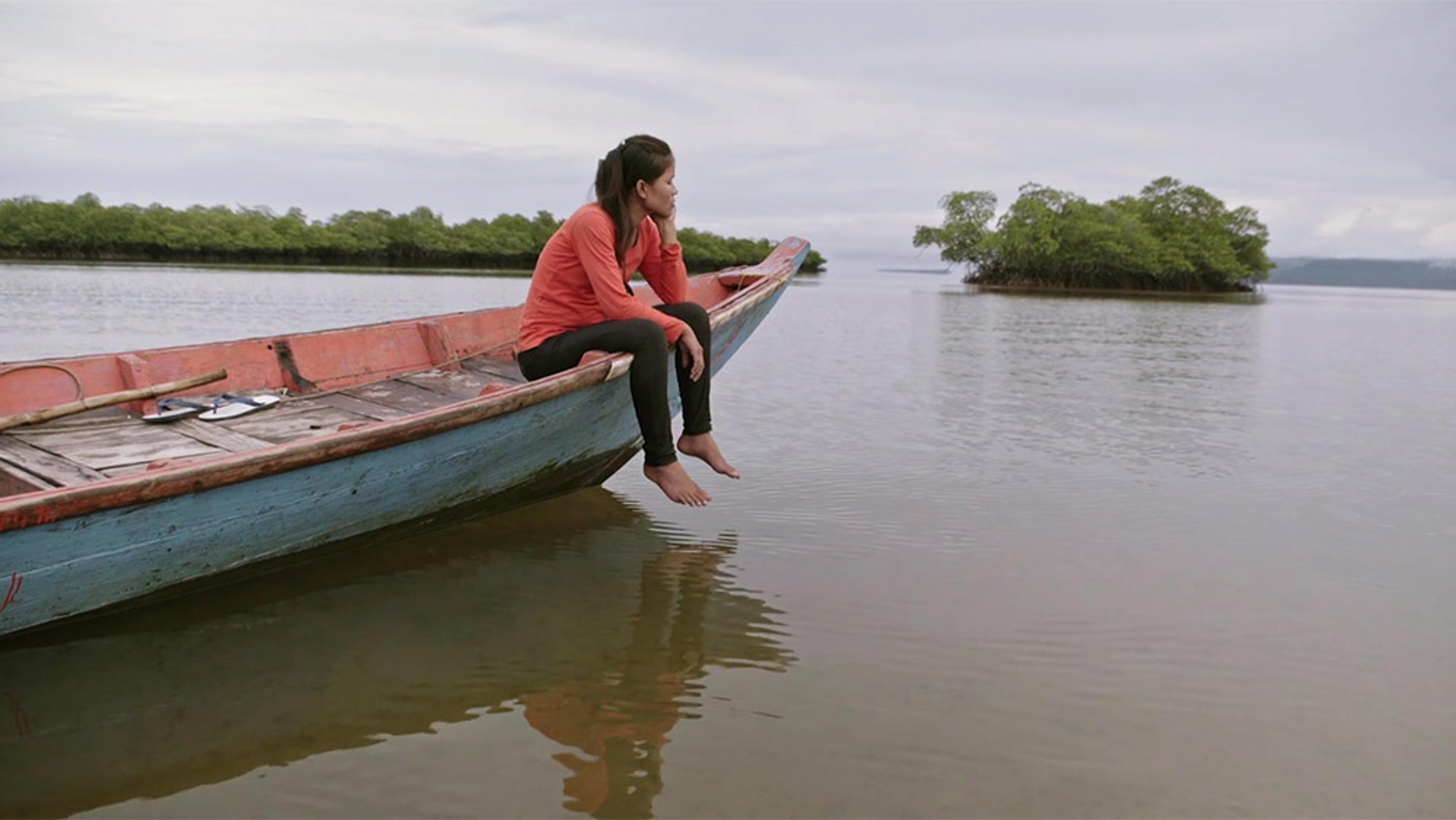 A woman sitting on the edge of a wooden boat in calm waters, with a view of distant islands and overcast sky.