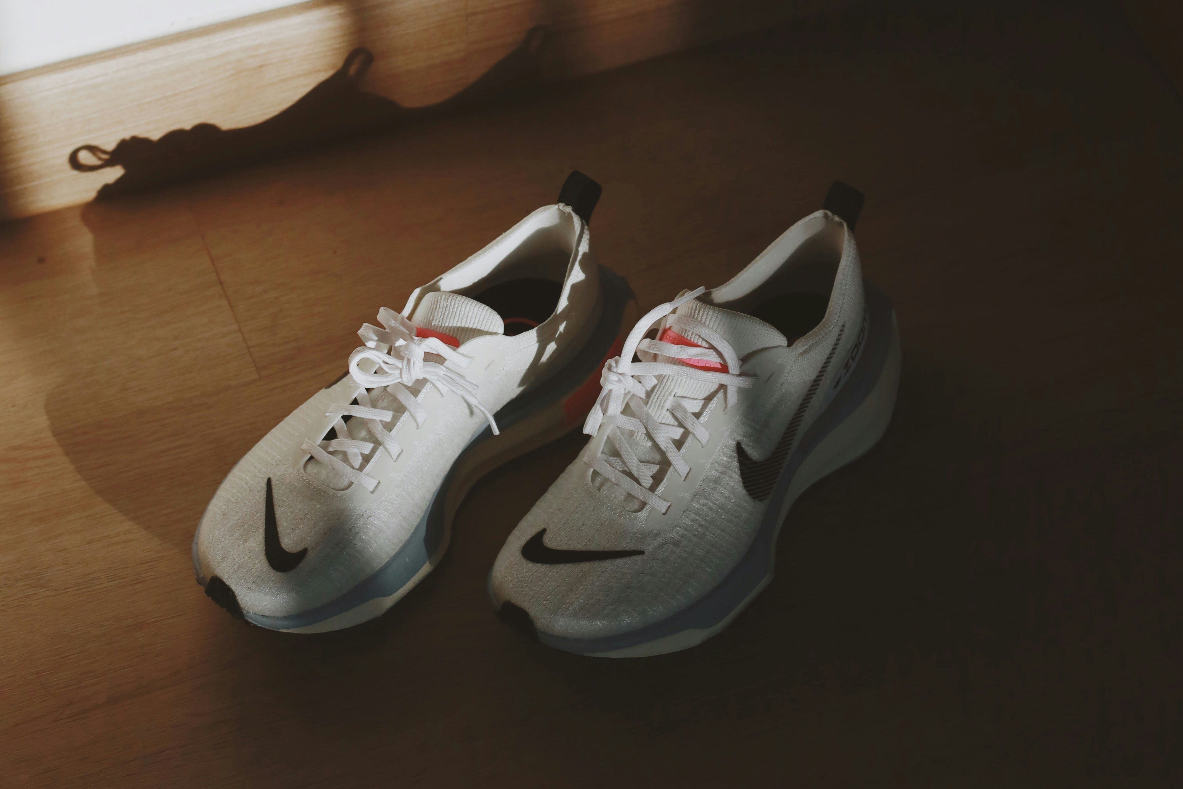 A pair of white trainers with a black tick Nike logo against a wooden floor, sunlight casting a shadow behind.
