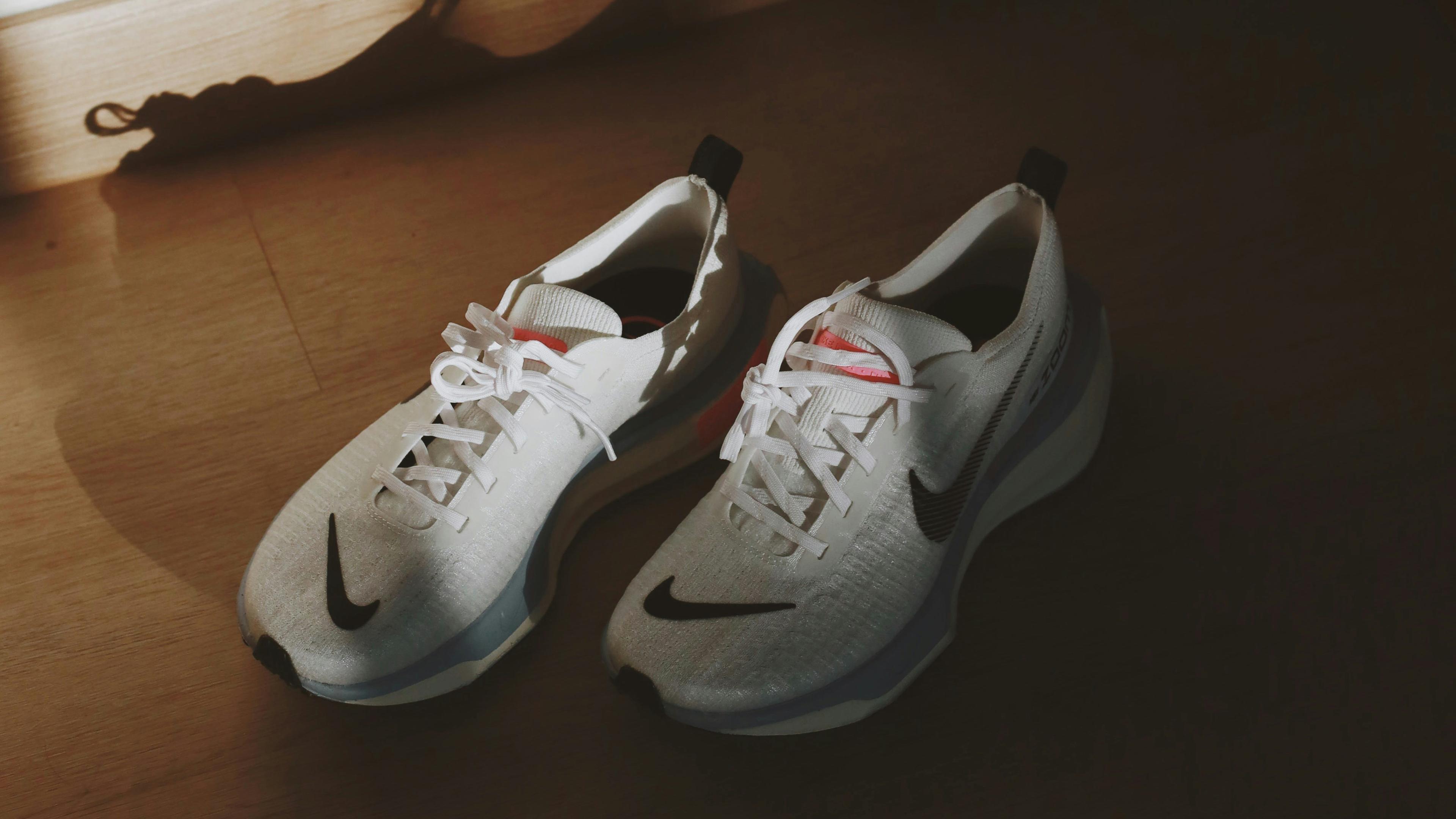 A pair of white trainers with a black tick Nike logo against a wooden floor, sunlight casting a shadow behind.