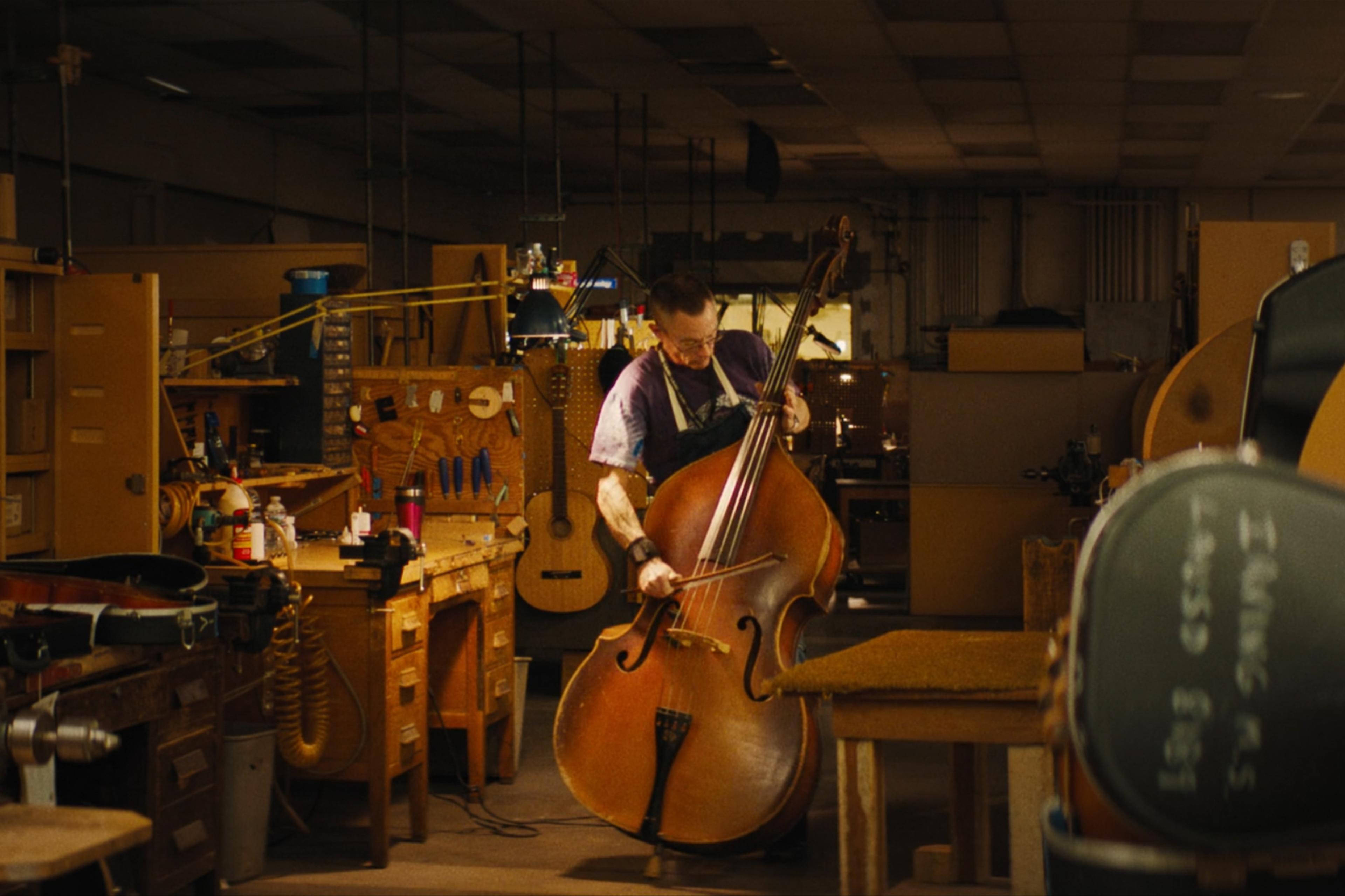 A person playing a double bass in a workshop filled with tools, workbenches, and various musical instruments.