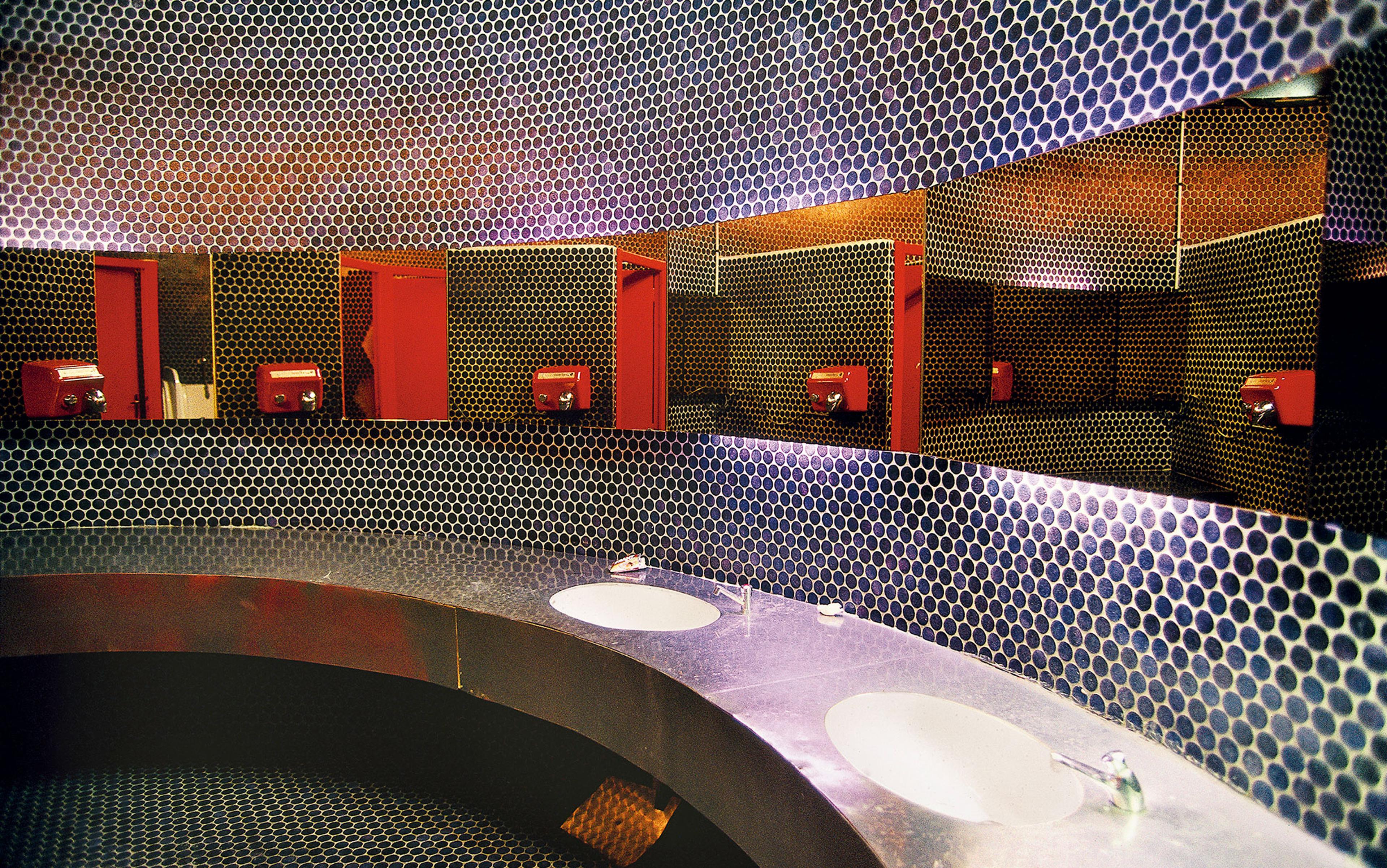 Photo of a modern bathroom with circular tiled walls and surfaces, two sinks, mirrors and red hand dryers reflected in the mirrors.