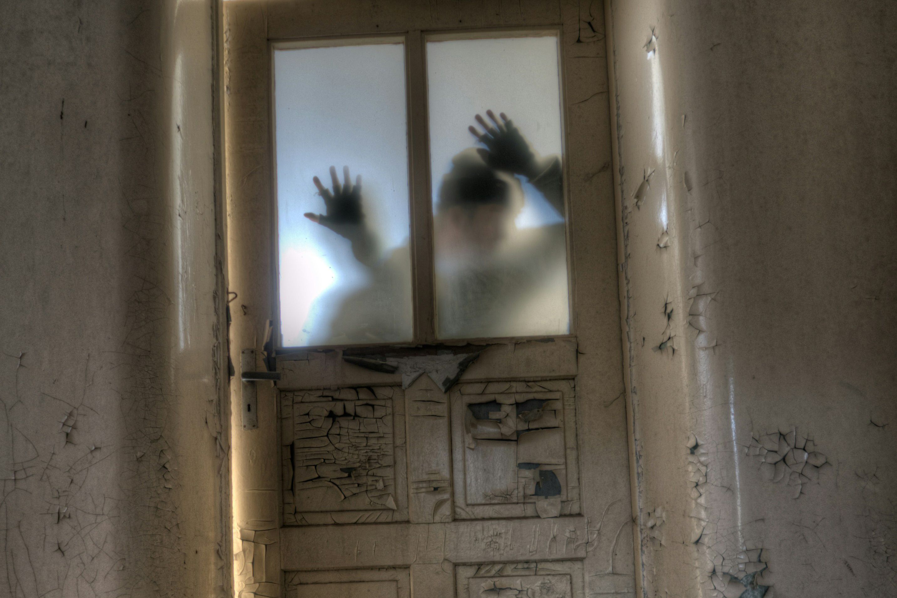 A silhouette pressing hands on a frosted glass door in an abandoned room with peeling paint on walls and door.