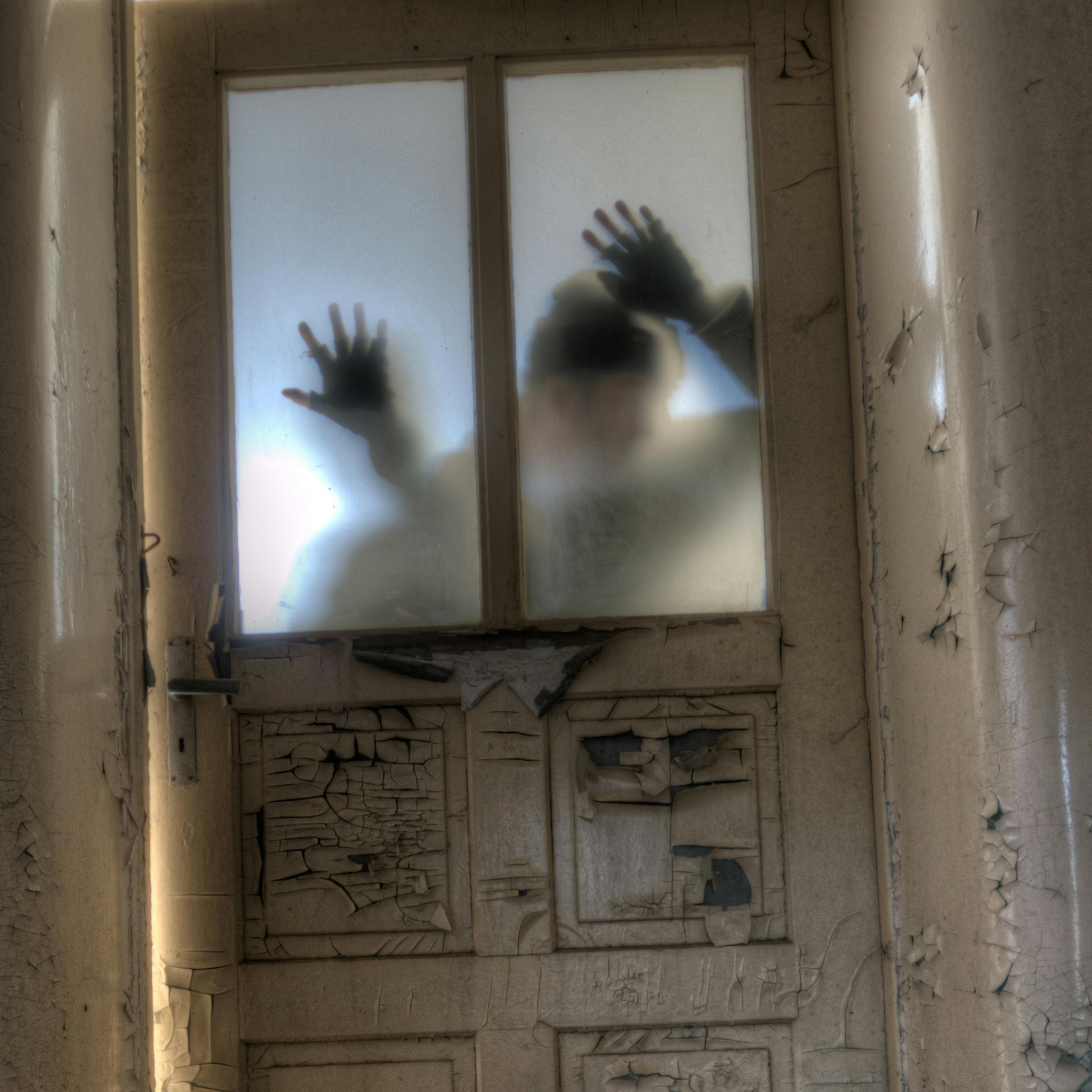 A silhouette pressing hands on a frosted glass door in an abandoned room with peeling paint on walls and door.