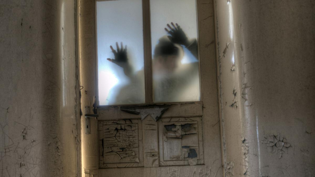 A silhouette pressing hands on a frosted glass door in an abandoned room with peeling paint on walls and door.