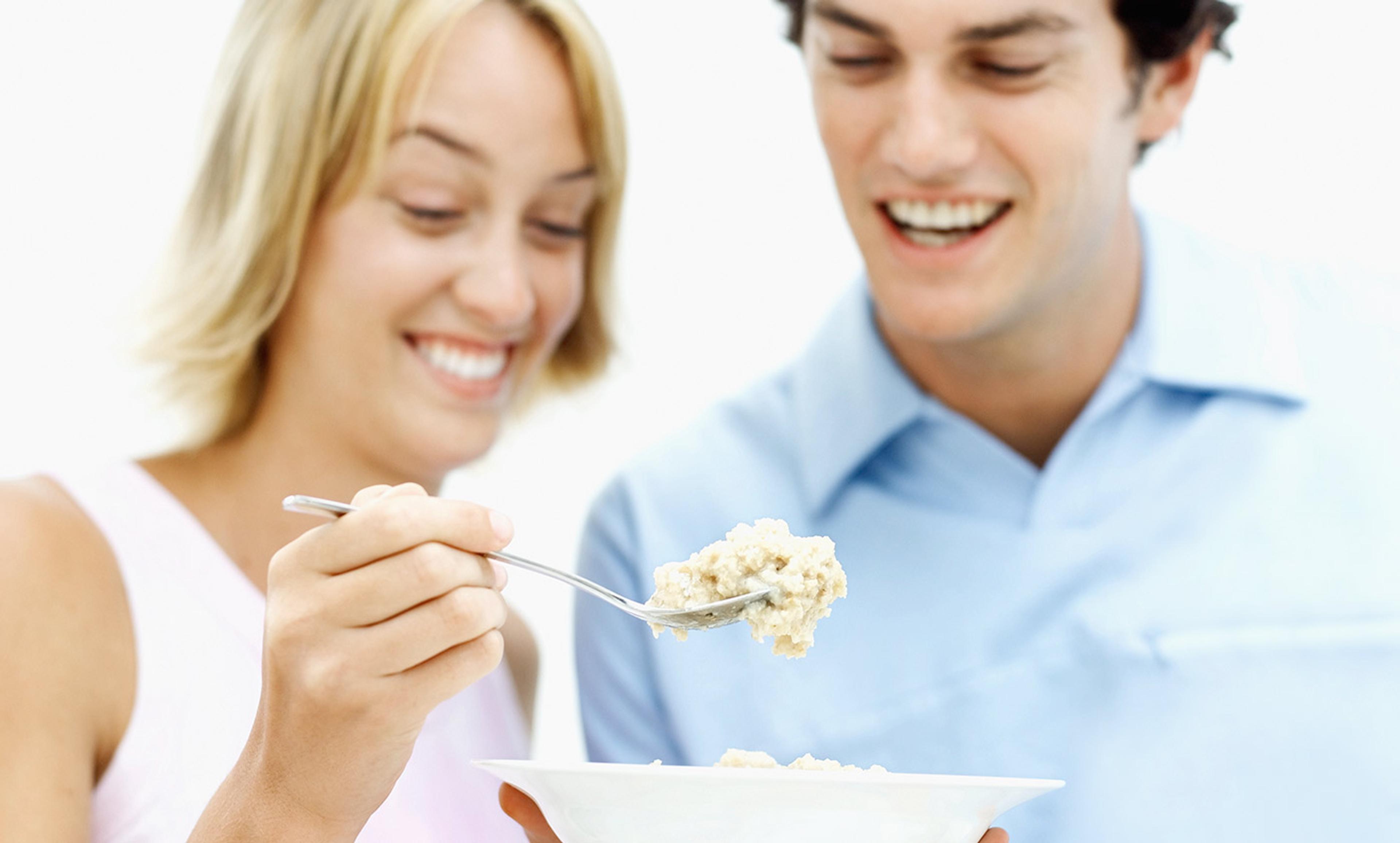 A smiling woman holding a spoonful of food, with a smiling man standing close beside her.