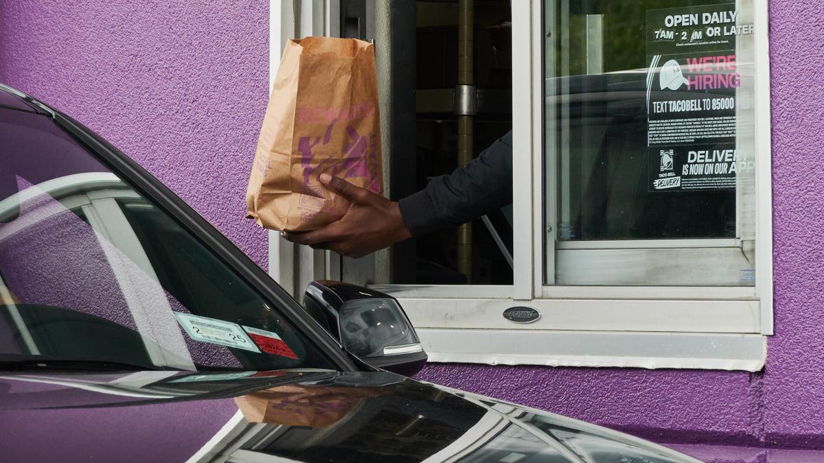 A drive-thru window with a hand passing a paper bag to a car by a purple wall.