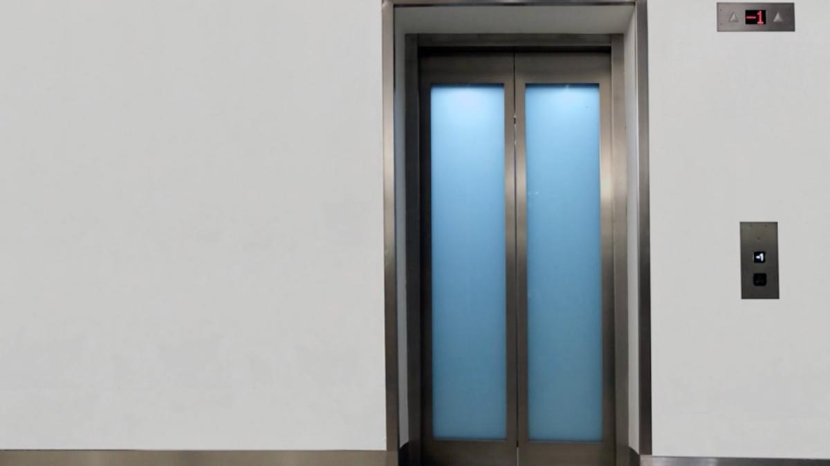 A closed lift with stainless steel doors and a blue panel display showing floor -1 above the button against a white wall.