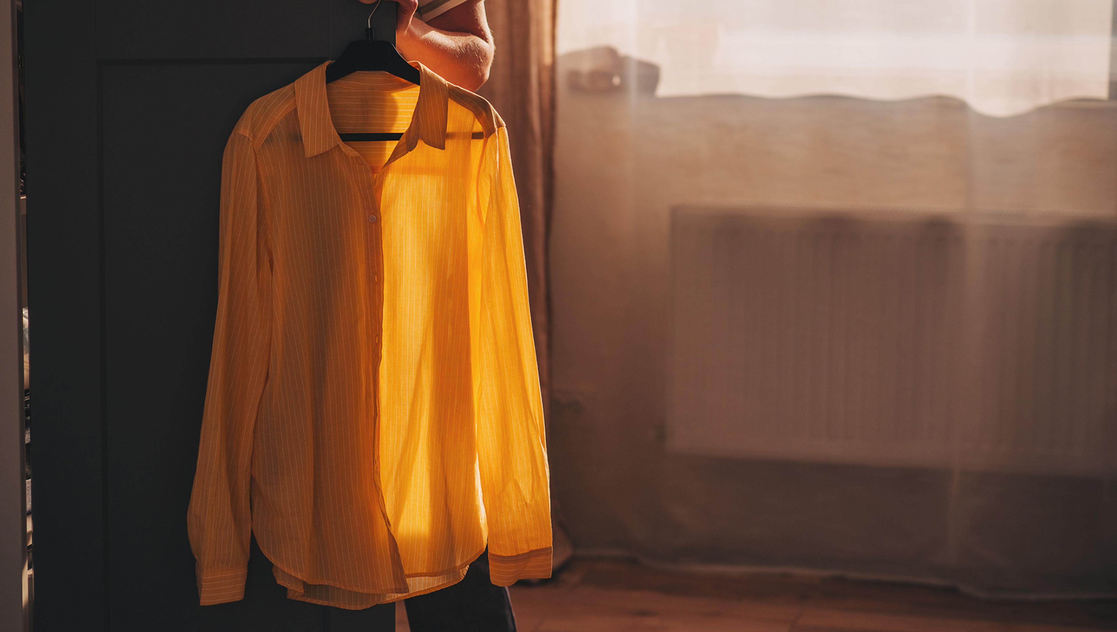 Photo of a yellow shirt on a hanger in soft sunlight with shadows on a dark cupboard door and a radiator in the background behind a sheer curtain. A person is holding the cupboard door open; their left arm is shown along with the sleeve of their striped t-shirt.