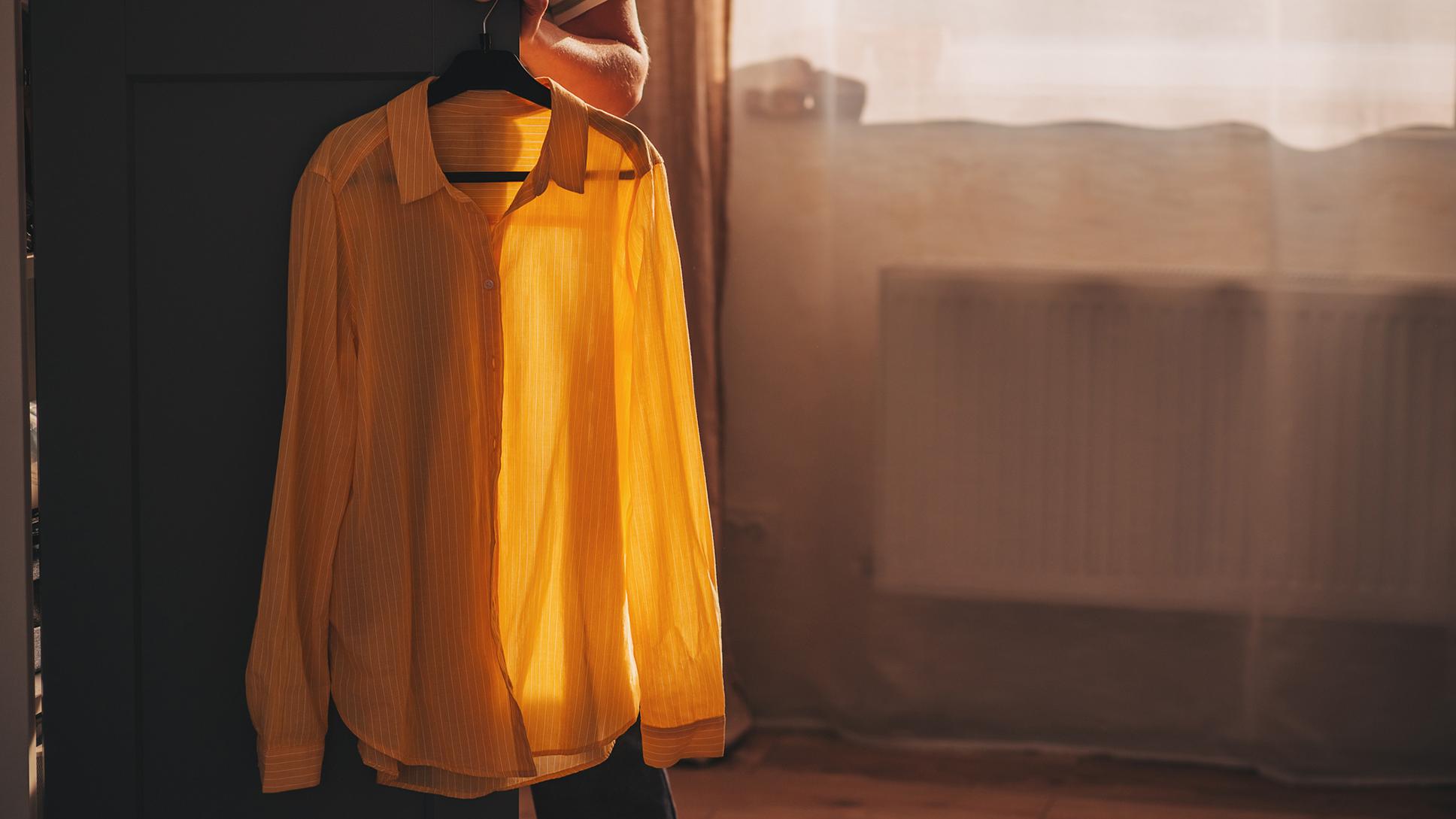 Photo of a yellow shirt on a hanger in soft sunlight with shadows on a dark cupboard door and a radiator in the background behind a sheer curtain. A person is holding the cupboard door open; their left arm is shown along with the sleeve of their striped t-shirt.