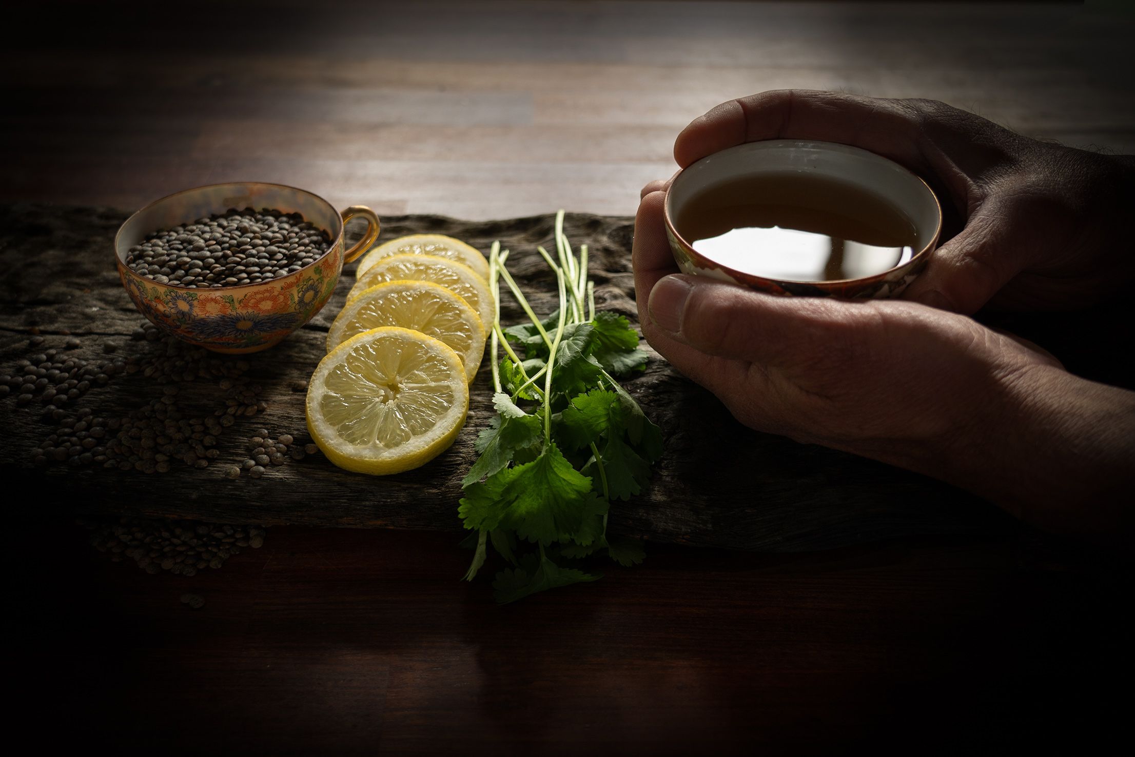 Hands holding tea, with a cup of lentils, lemon slices and coriander on a wooden board in dim light.