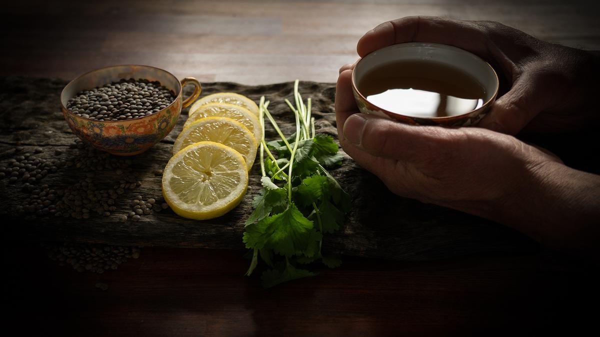 Hands holding tea, with a cup of lentils, lemon slices and coriander on a wooden board in dim light.