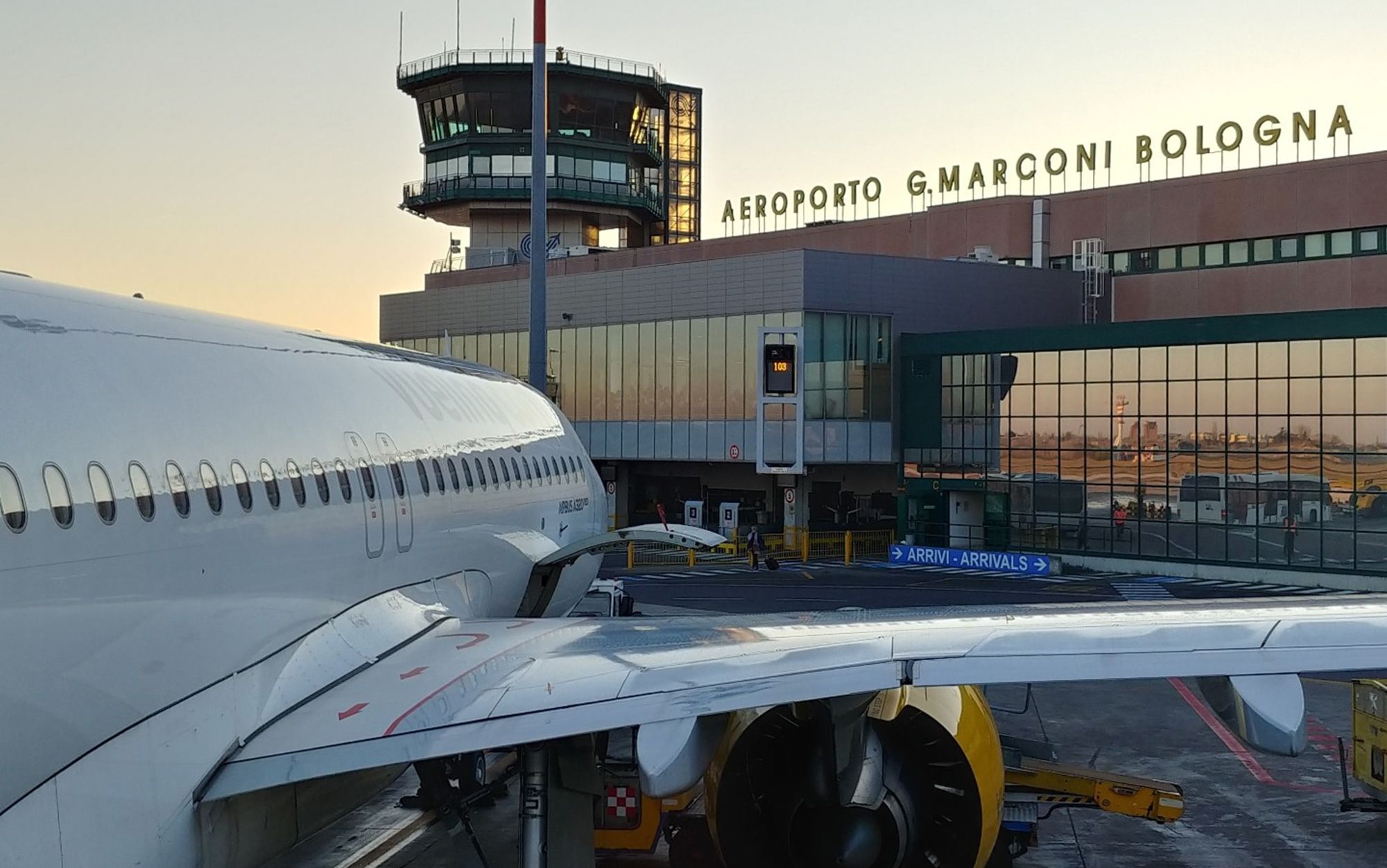 A plane docked at Bologna Airport at sunset with the terminal and control tower in the background.