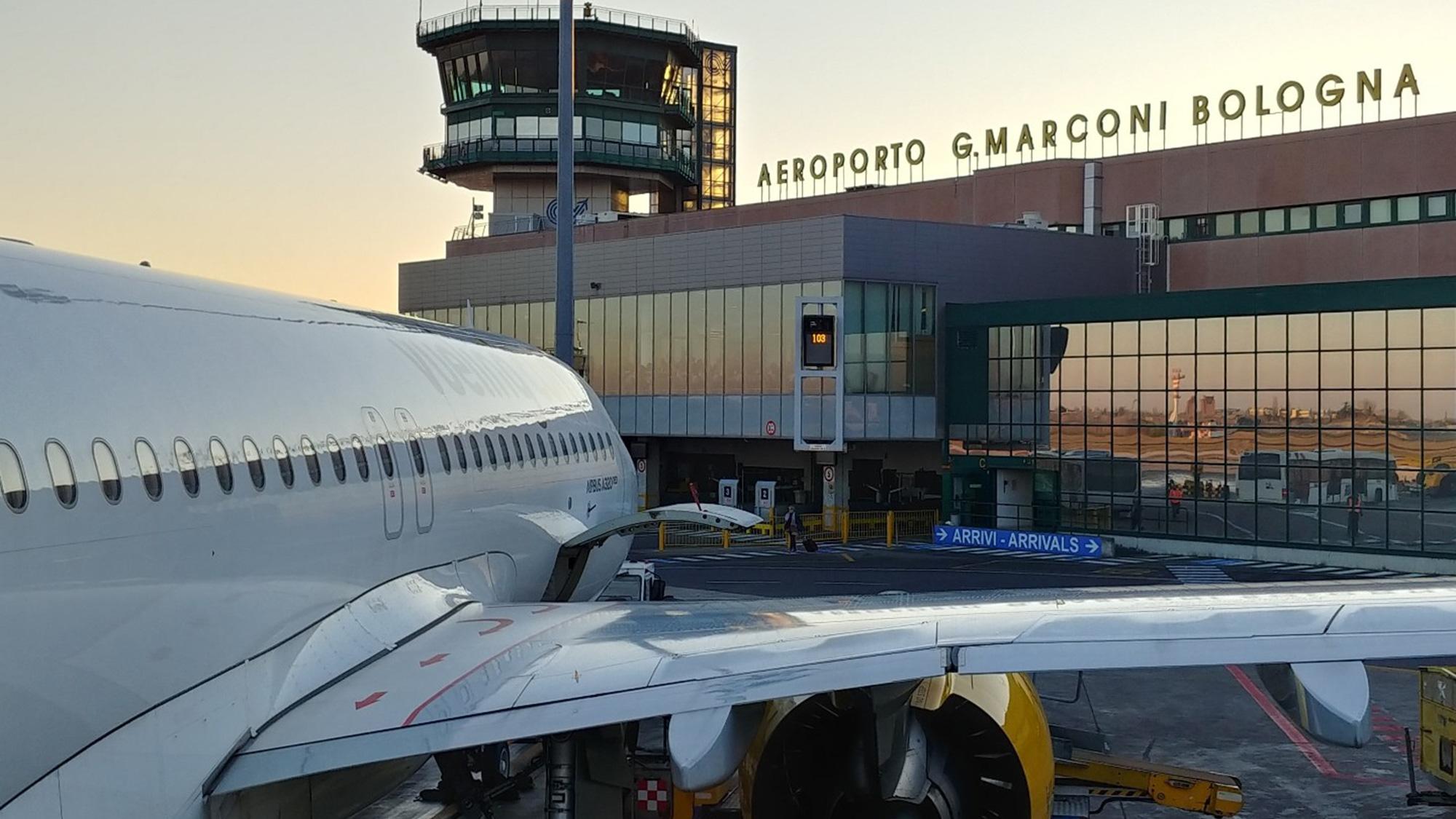 A plane docked at Bologna Airport at sunset with the terminal and control tower in the background.