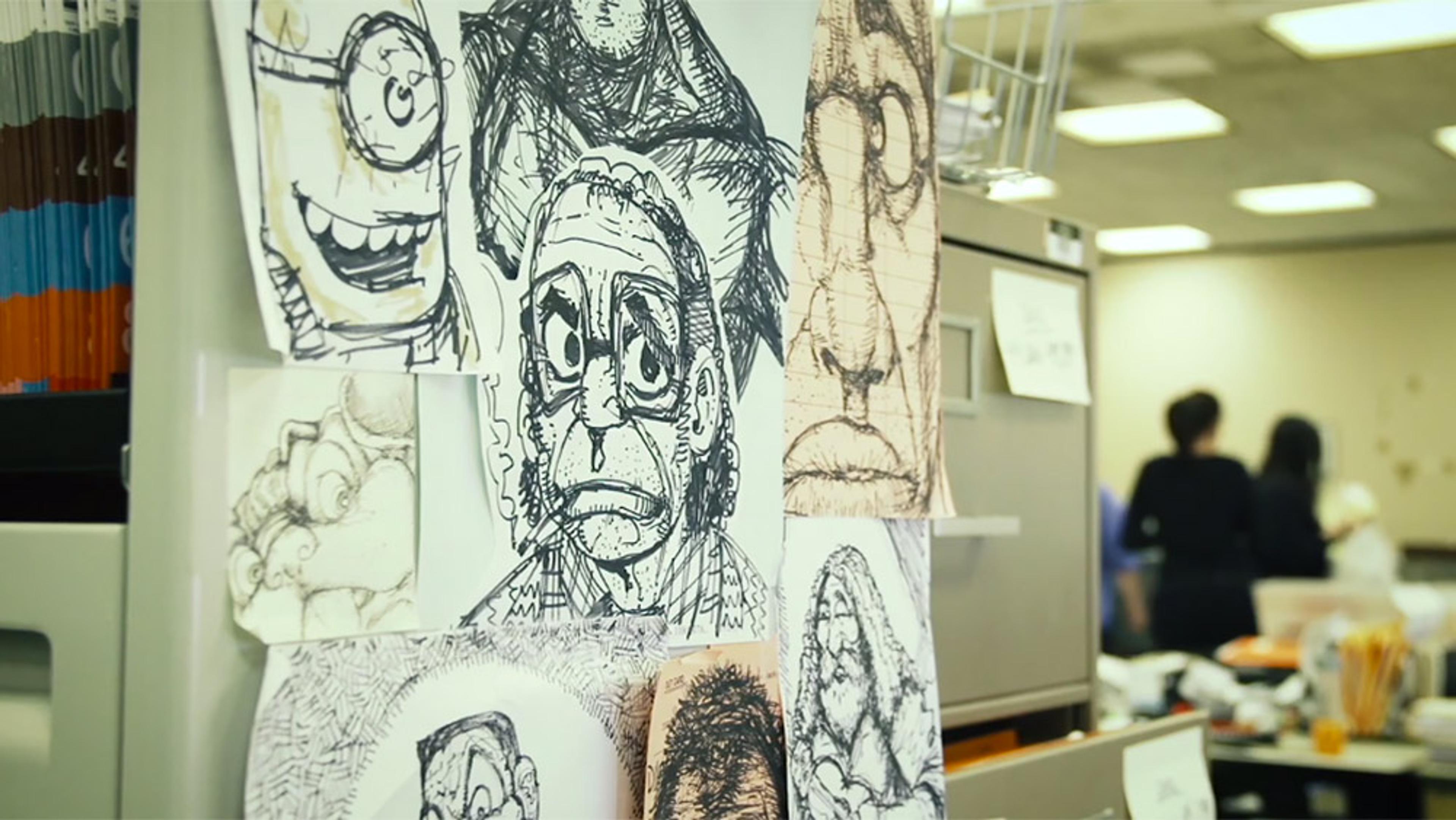 Photo of cartoon sketches pinned to an office wall, with people working in the background under fluorescent lights.