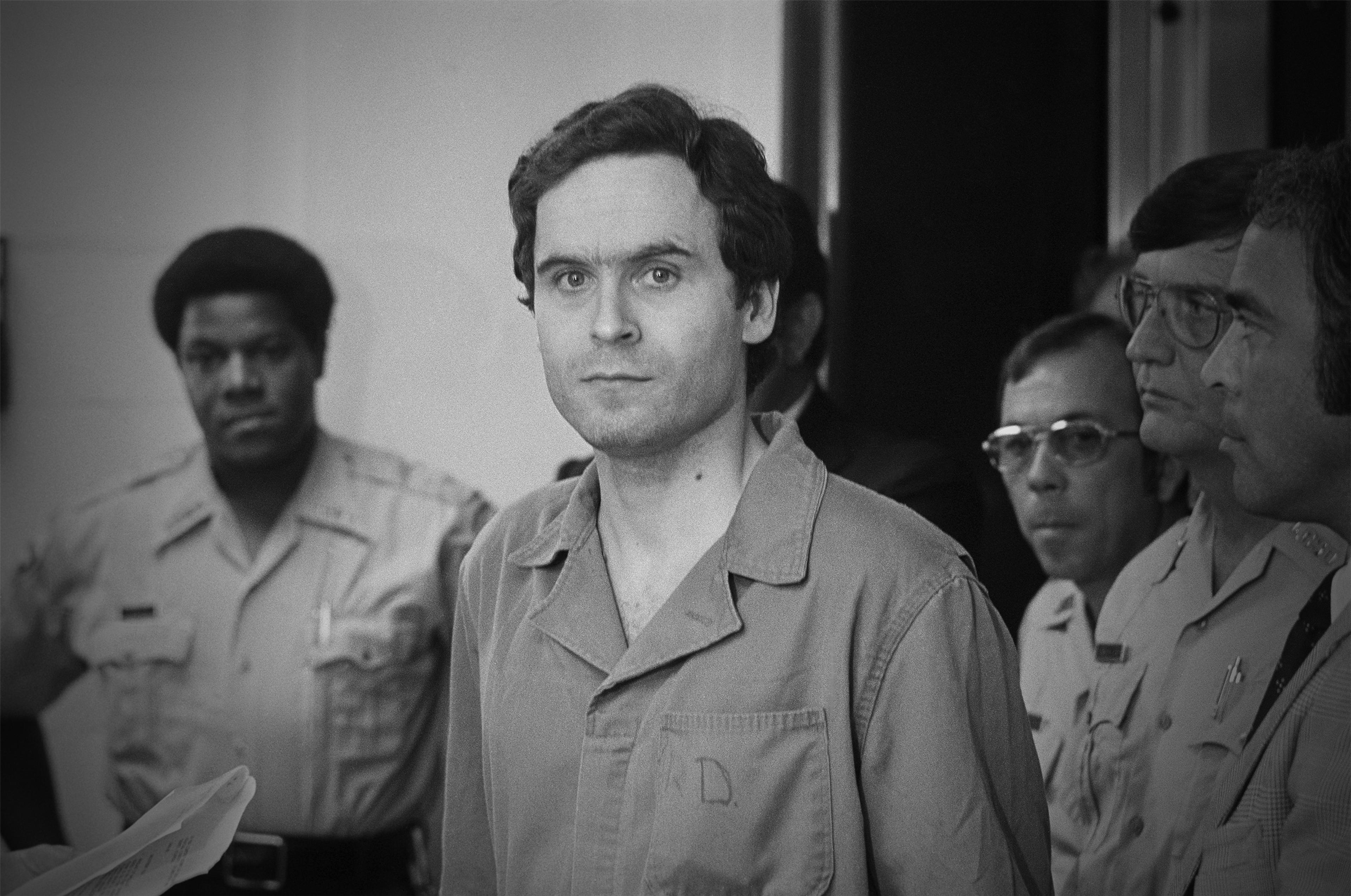 Black and white photo of a man in a prison uniform surrounded by security personnel in a courtroom setting.