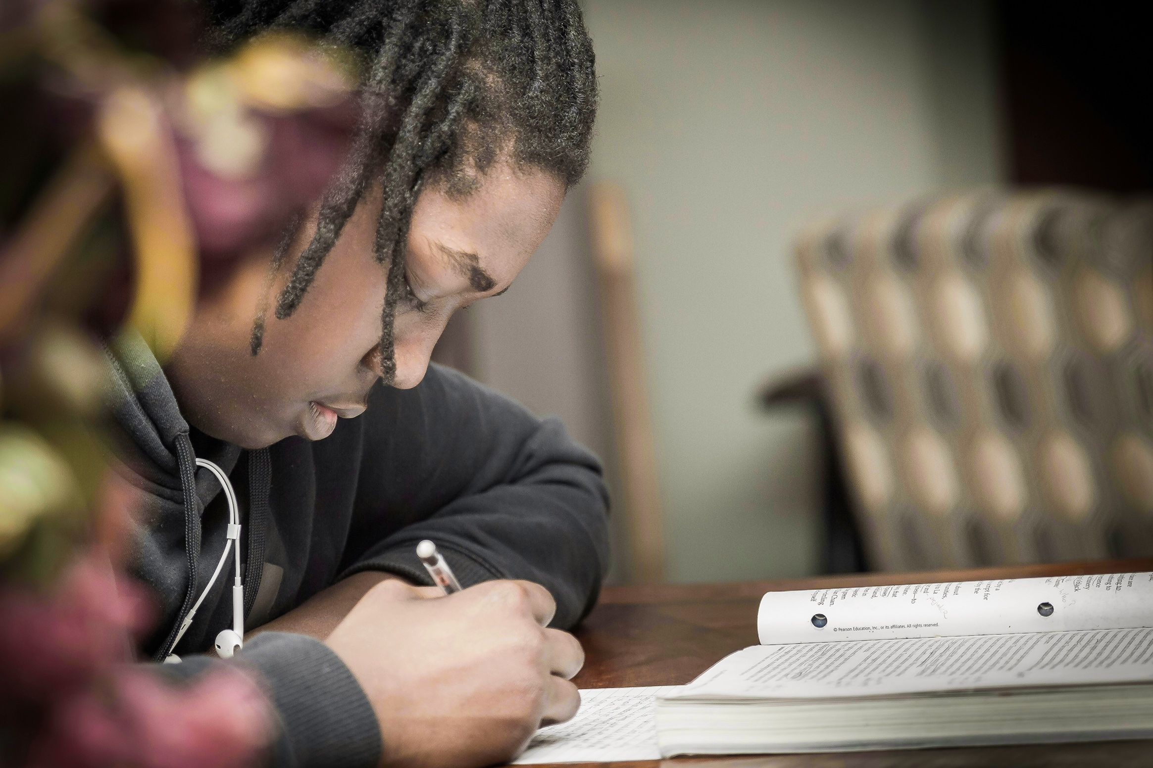 A child writing in a notebook at a table, wearing a dark hoodie, with blurred background.
