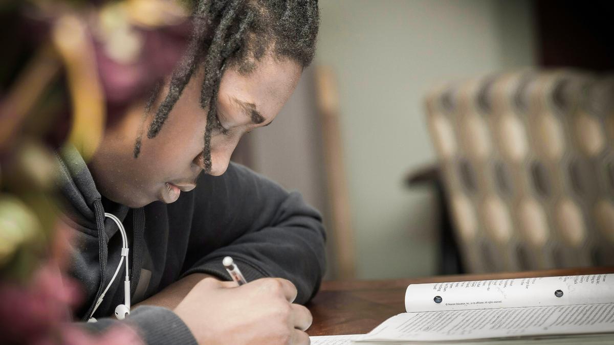 A child writing in a notebook at a table, wearing a dark hoodie, with blurred background.
