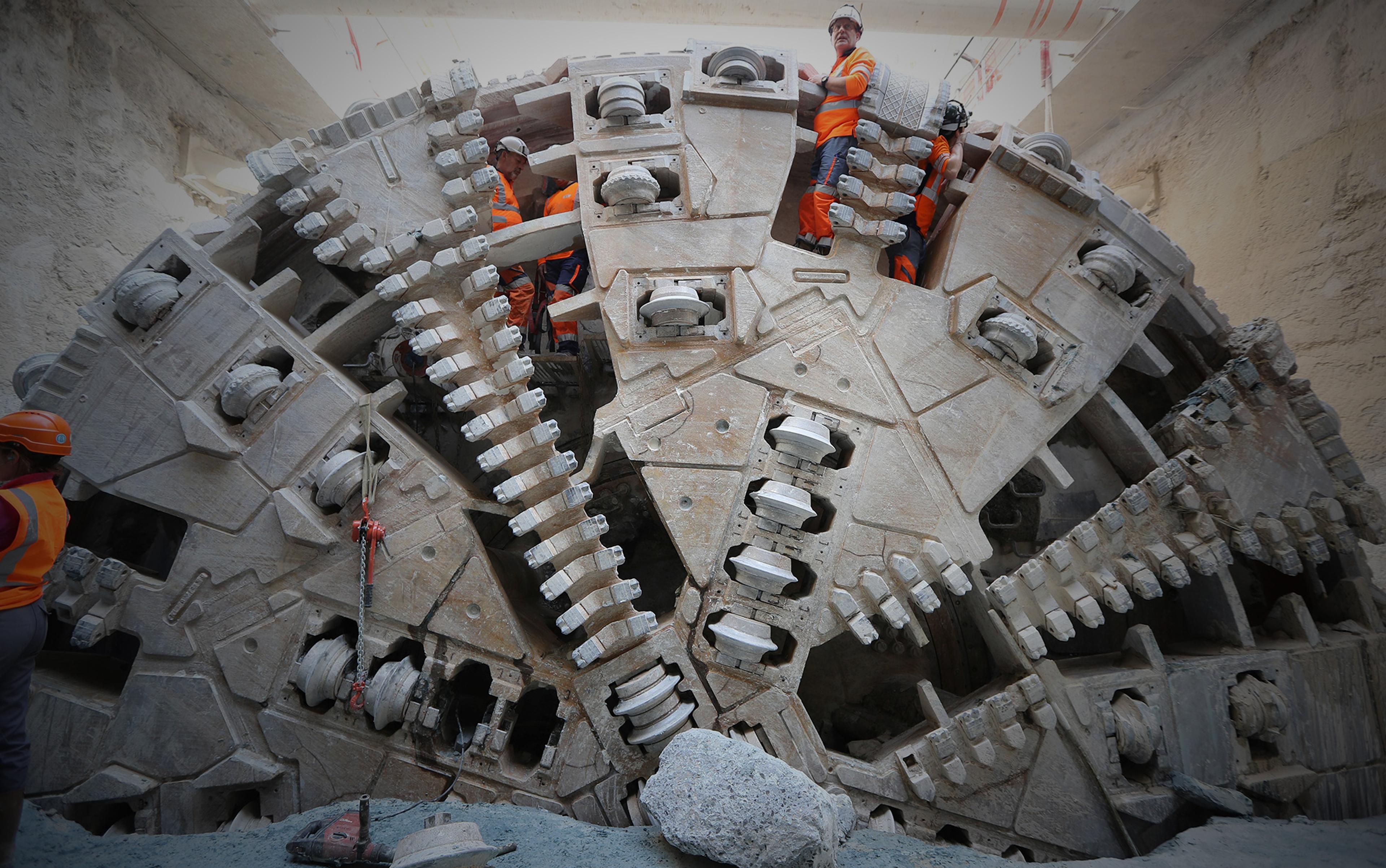 Construction workers standing on an enormous tunnelling machine in an underground site, wearing orange safety vests and helmets.