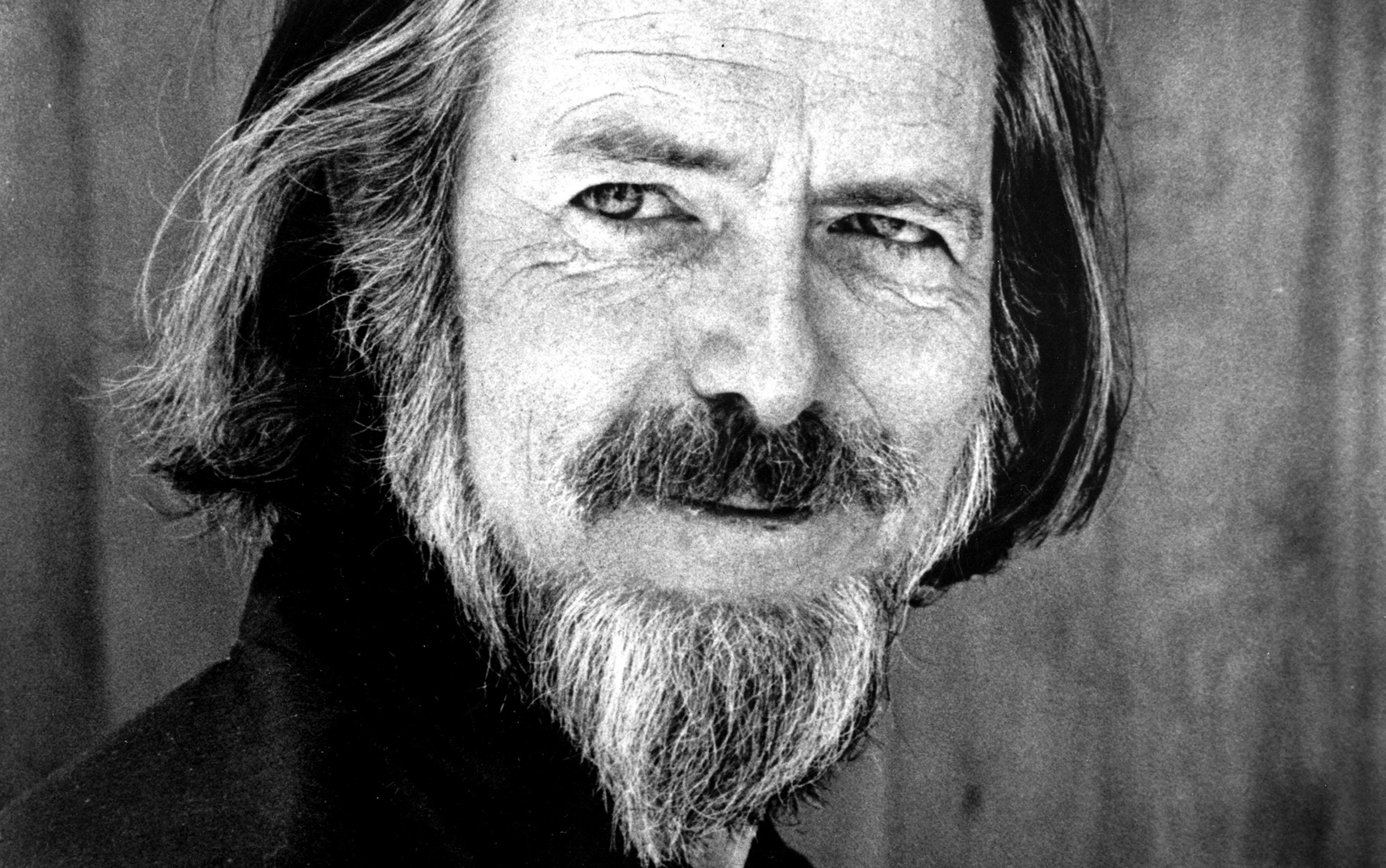 Close-up of a man with a long beard and shoulder-length hair, smiling slightly with a contemplative expression, black and white photograph.