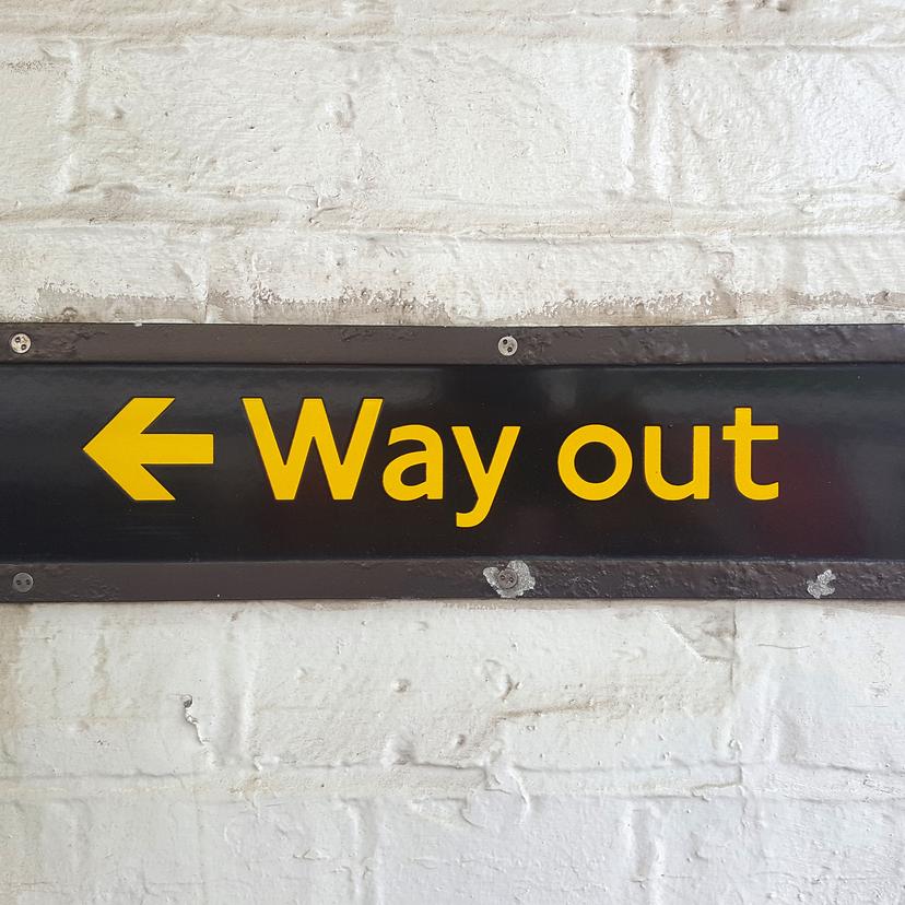 Photo of a black sign with yellow text reading ‘Way out’ and a left-pointing arrow, mounted on a white brick wall.