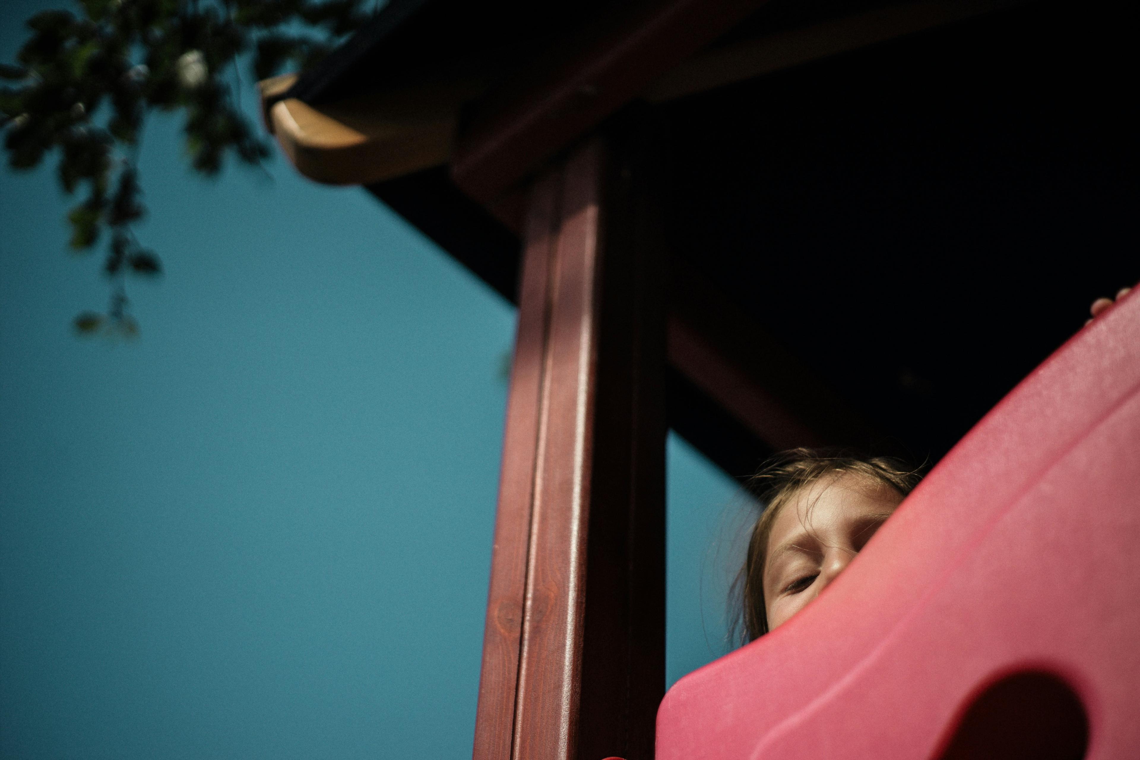Photo of a child peeking over a pink slide against a blue sky, partially obscured by play equipment and tree branches.