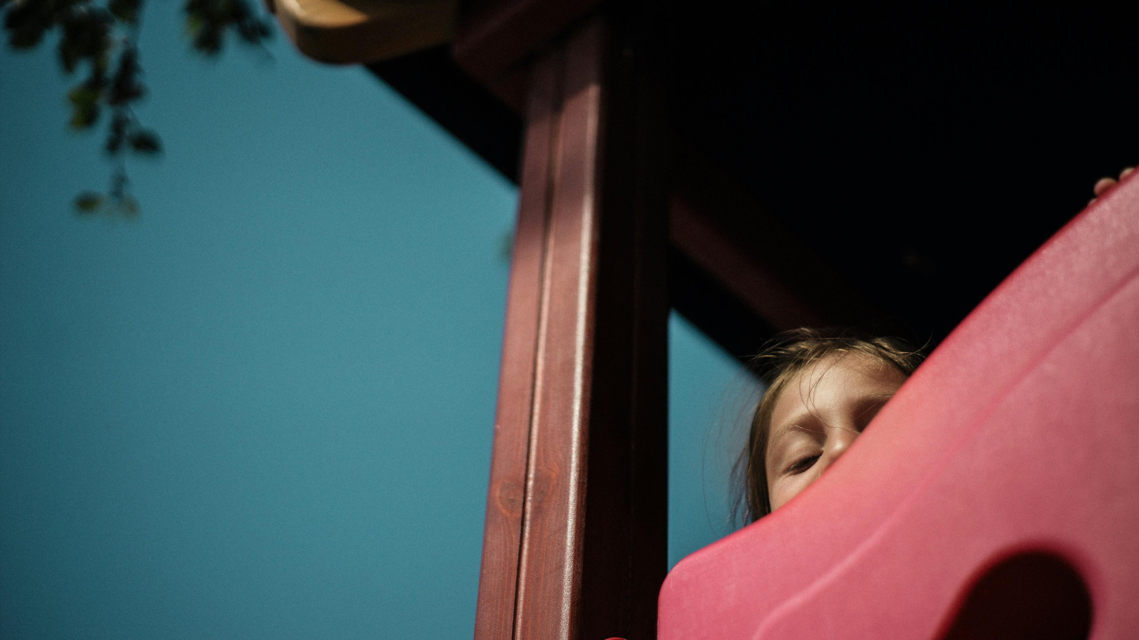 Photo of a child peeking over a pink slide against a blue sky, partially obscured by play equipment and tree branches.