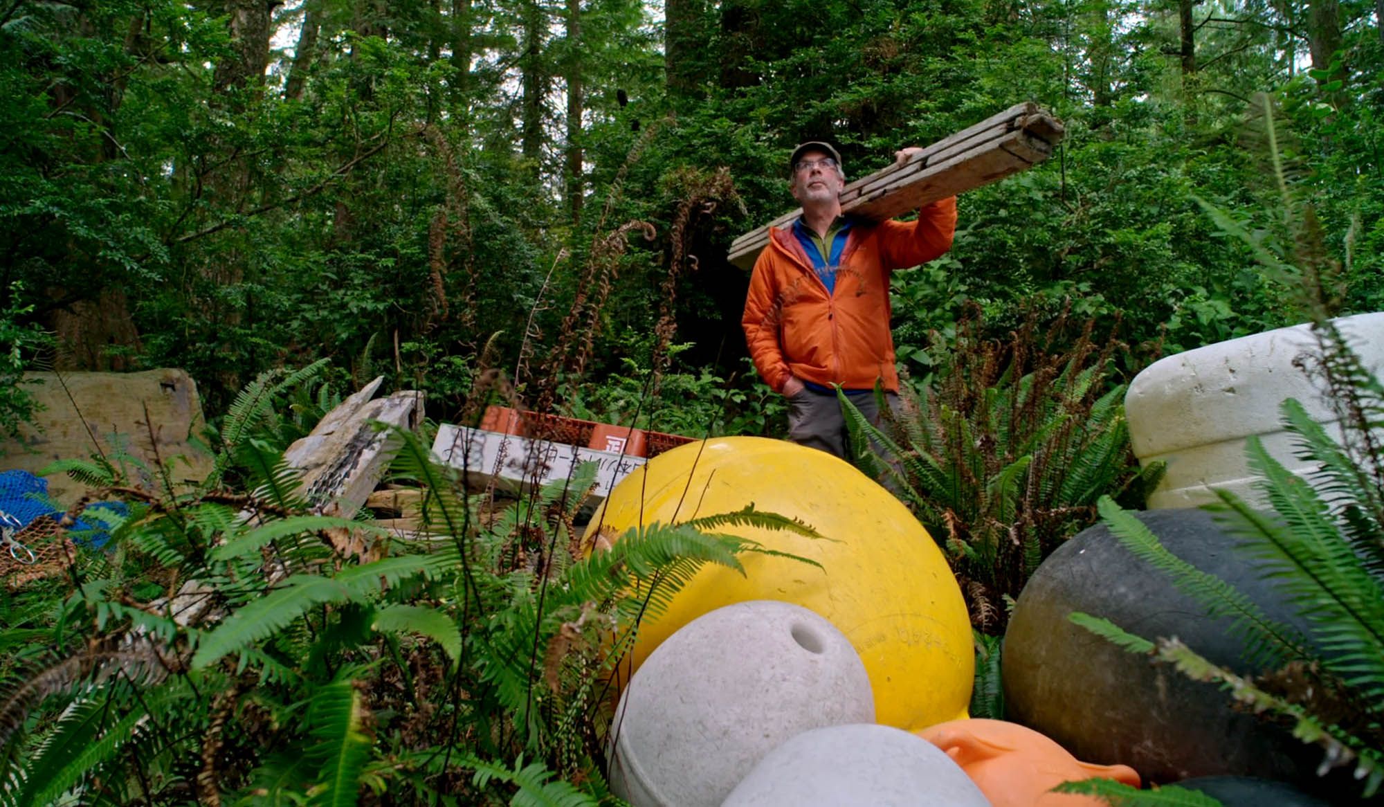 Man in an orange jacket carrying planks stands in a dense forest next to large colourful buoys and scattered debris.