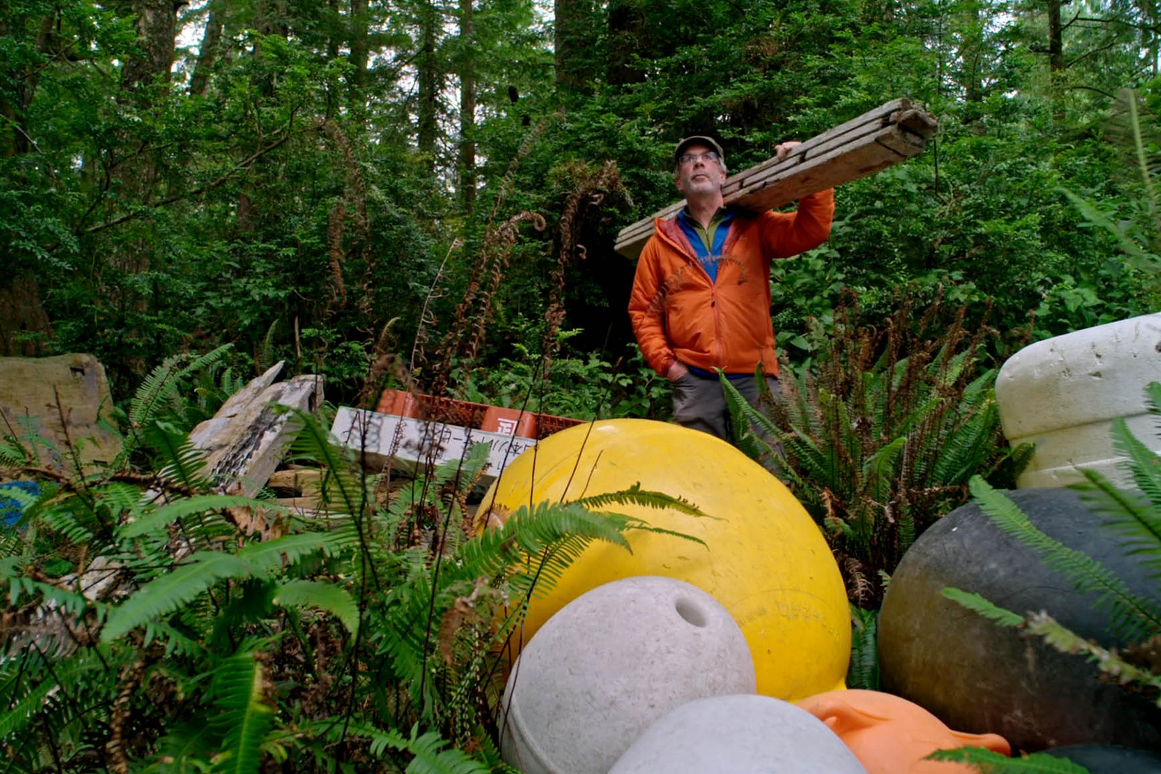 Man in an orange jacket carrying planks stands in a dense forest next to large colourful buoys and scattered debris.