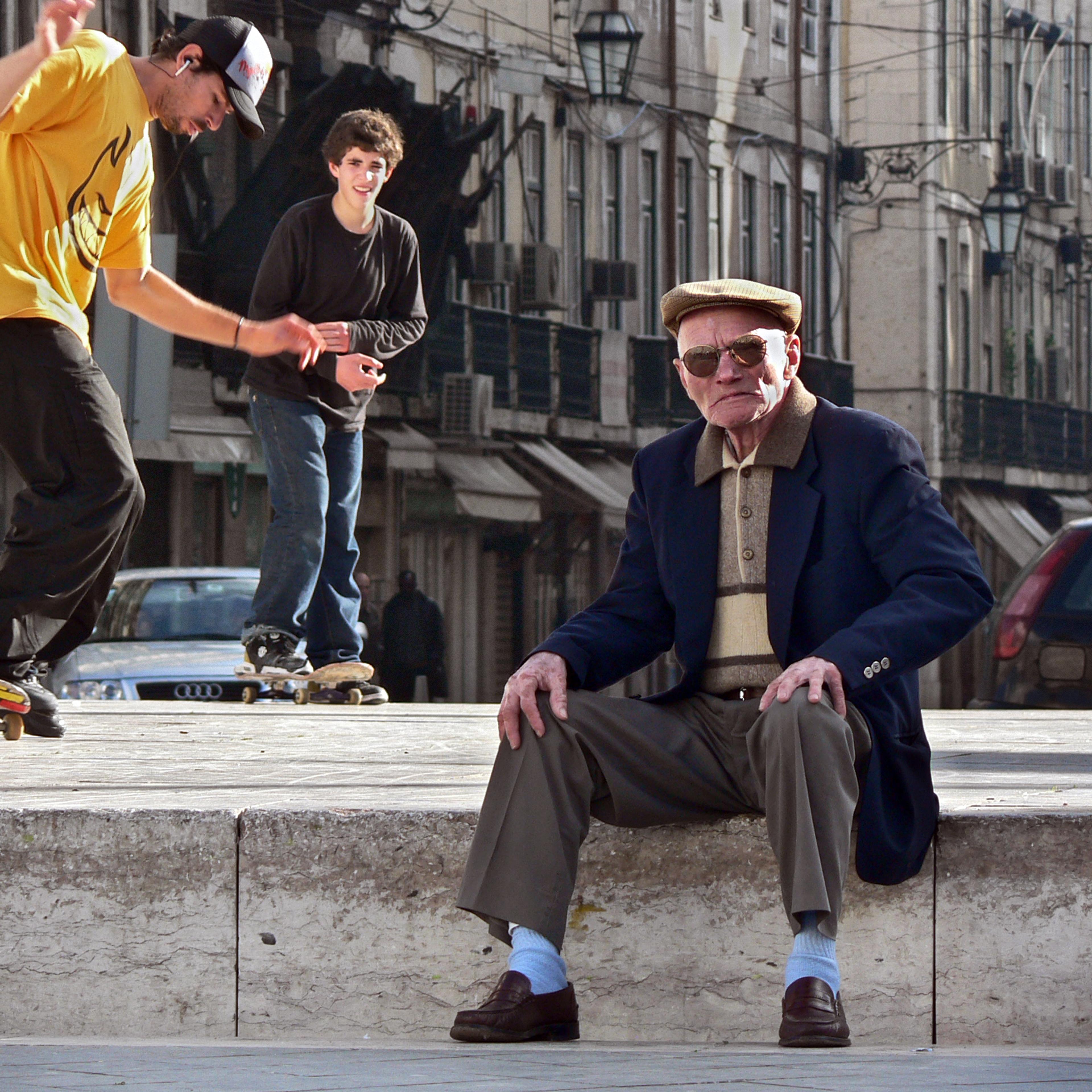 An elderly man seated on a city street as skateboarders perform tricks around him.