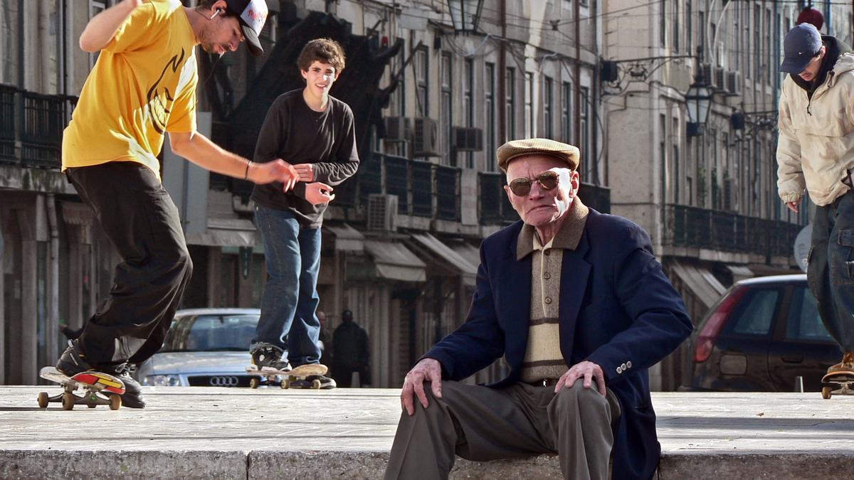 An elderly man seated on a city street as skateboarders perform tricks around him.