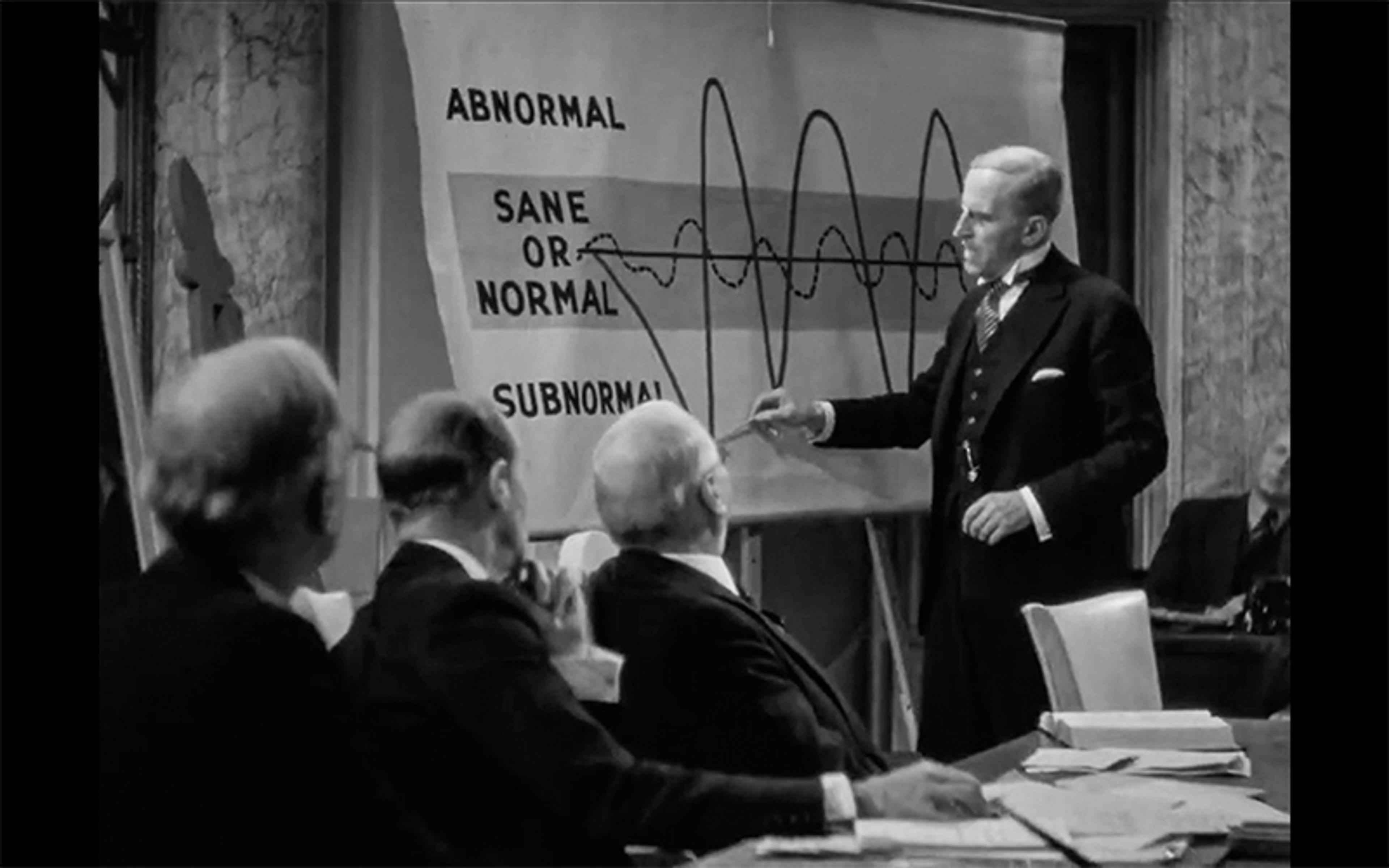 Black and white photo of a man presenting a chart to seated colleagues in an office setting, the chart has categories labelled ‘Abnormal’, ‘Sane or Normal’ and ‘Subnormal’.