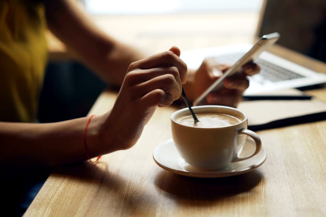 A person stirring coffee with a spoon while looking at a phone at a wooden table with a laptop in the background.