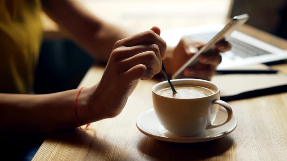 A person stirring coffee with a spoon while looking at a phone at a wooden table with a laptop in the background.