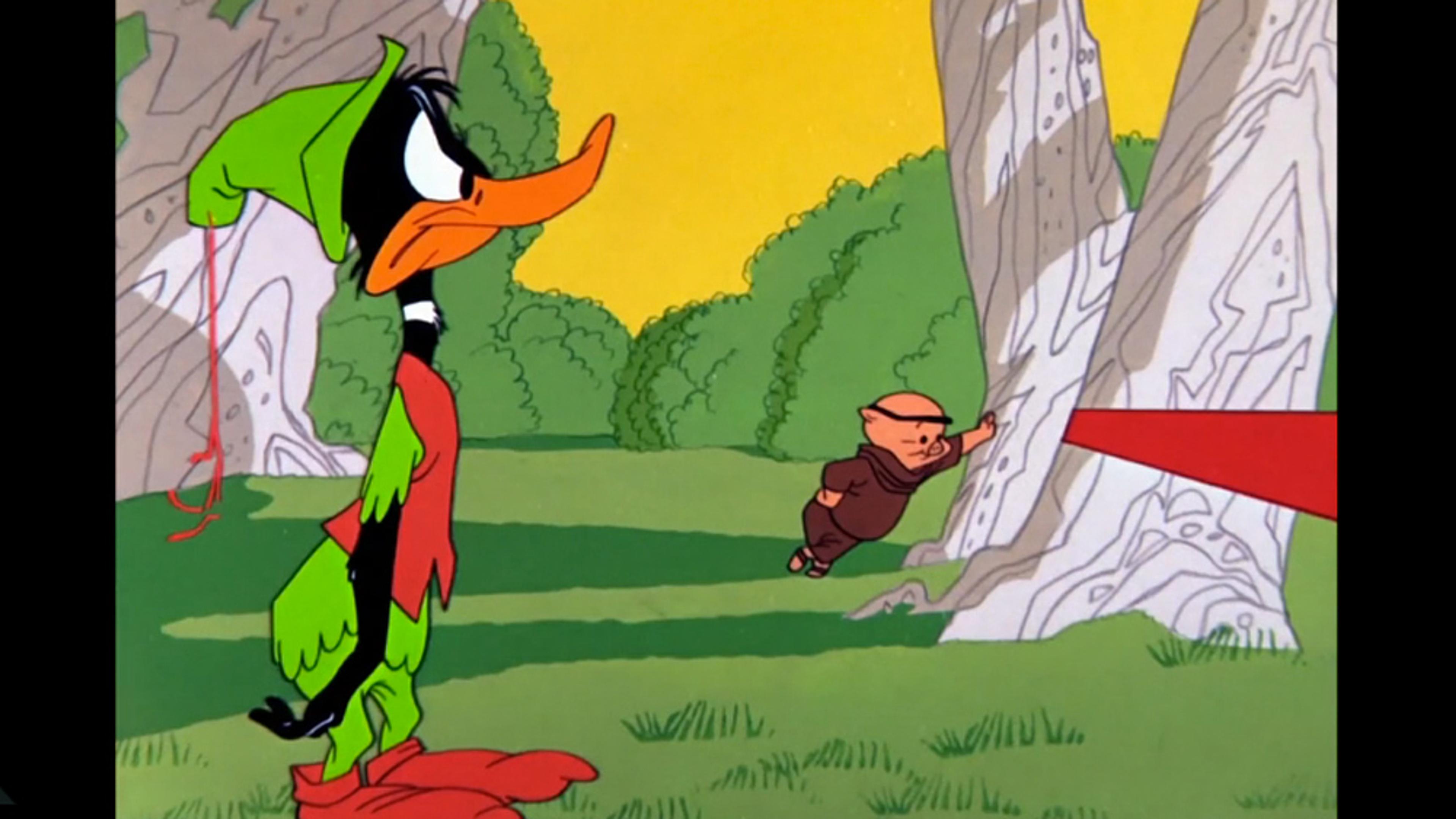 Animated Daffy Duck in a green outfit stands in a forest while a small man leans against a tree in the background.