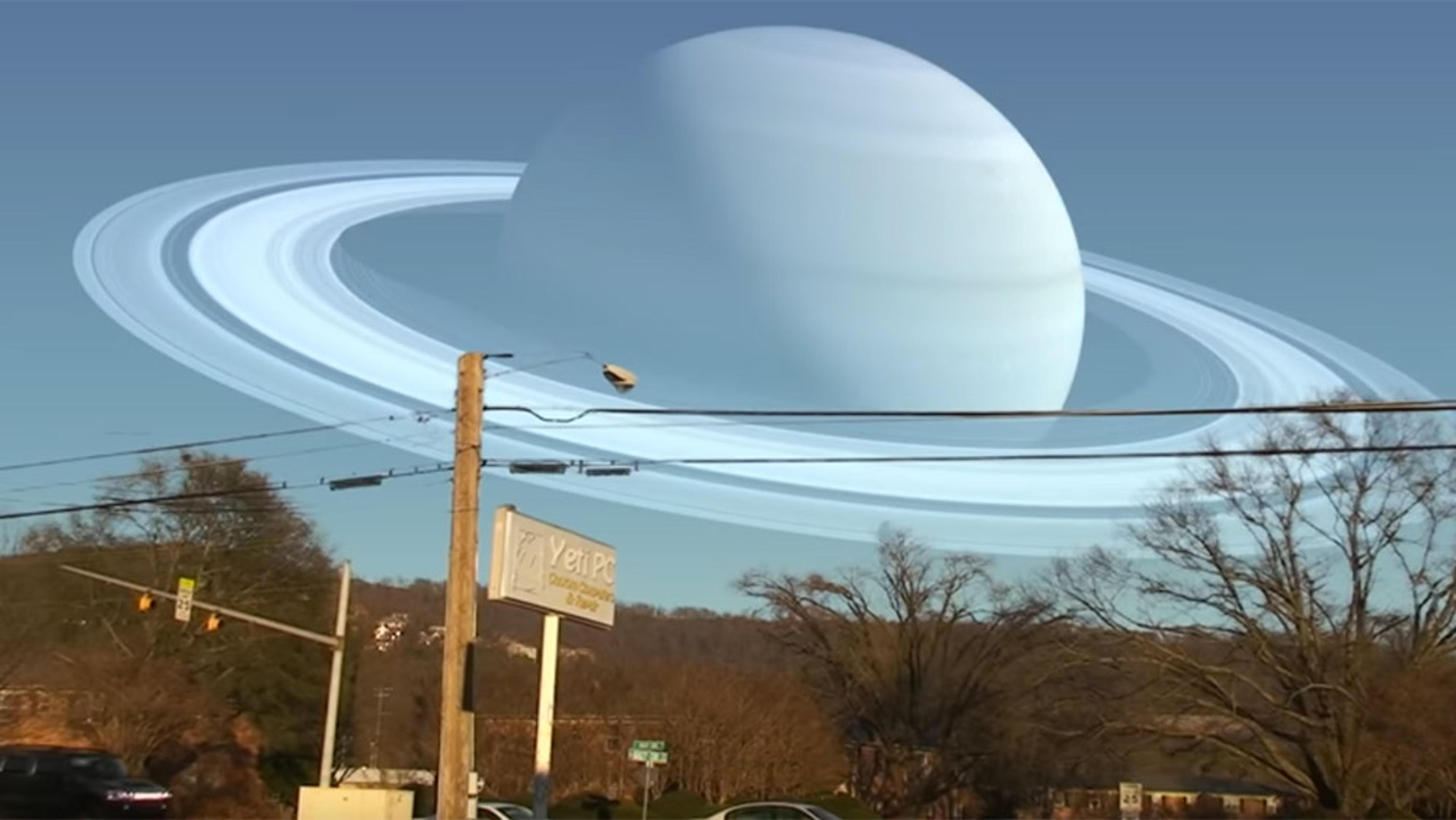 A street scene with a huge, digitally added Saturn dominating the sky, power lines, trees and a business sign visible.