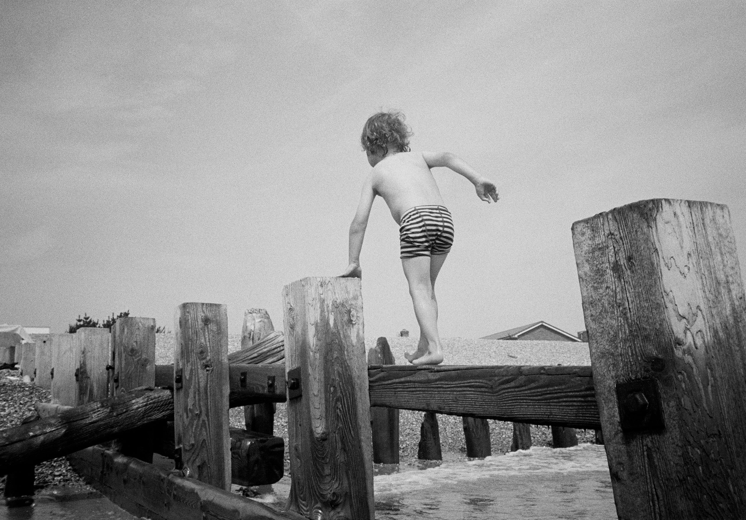 Black and white photo of a child in striped shorts balancing on wooden posts by the seaside.