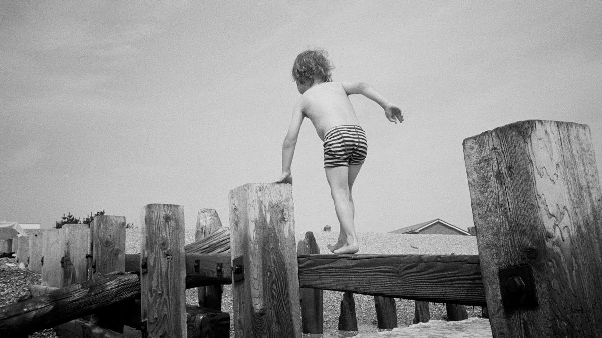 Black and white photo of a child in striped shorts balancing on wooden posts by the seaside.