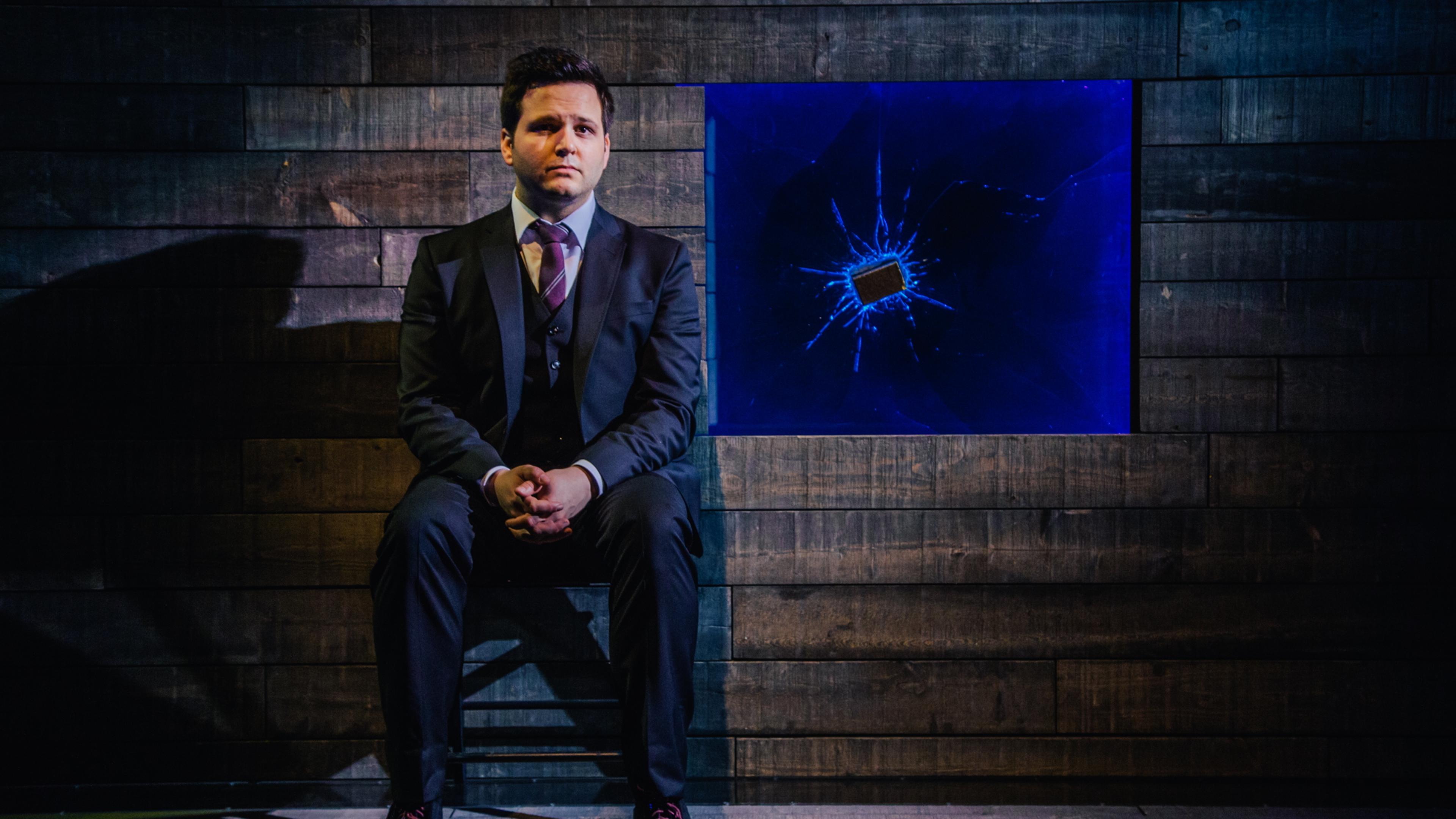 Photo of a man in a suit sitting in a dimly lit room with a shattered blue screen on a wooden wall behind him.
