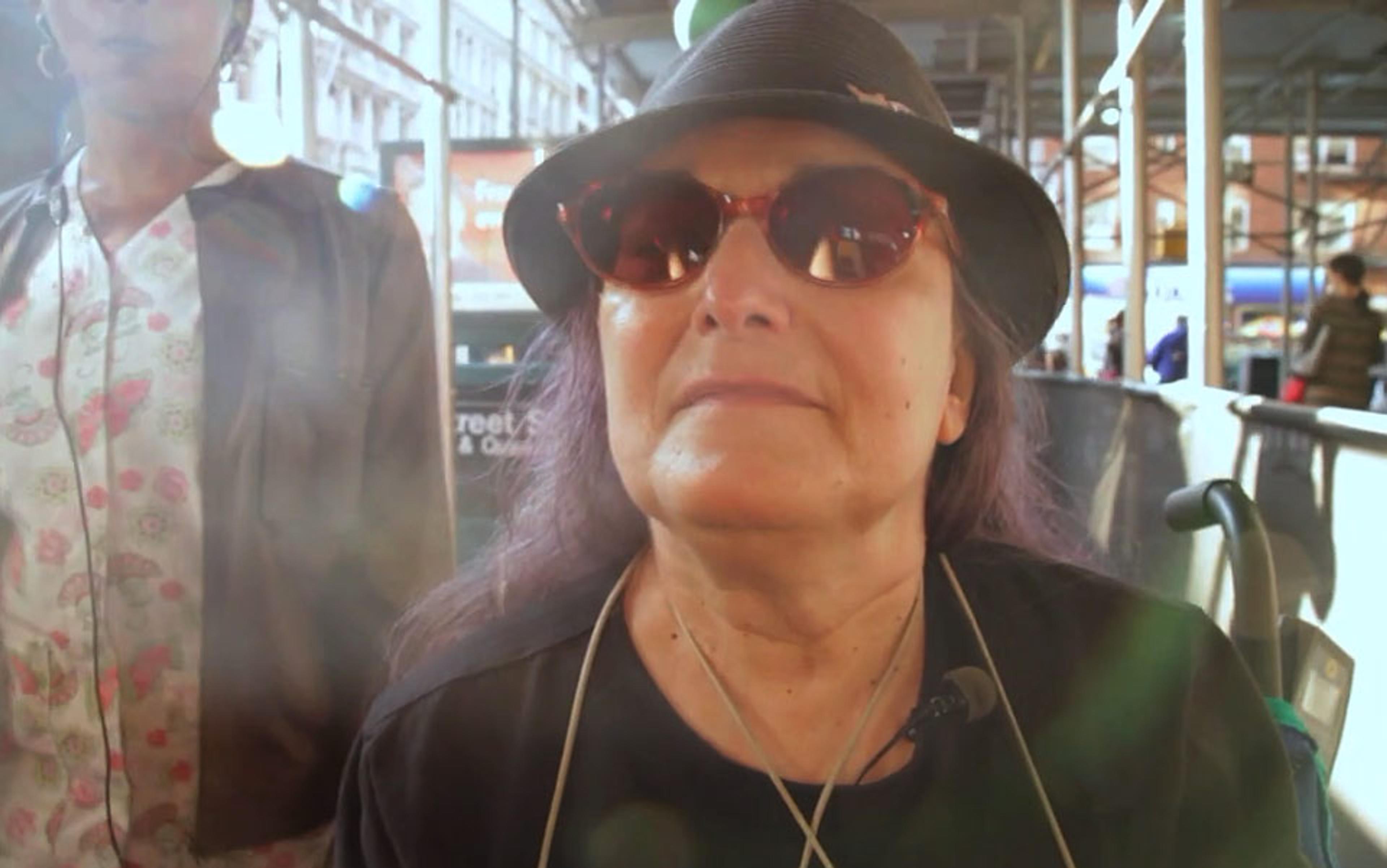 A woman with sunglasses, a hat and long purple hair in an outdoor setting. Another person is in the background with scaffolding.