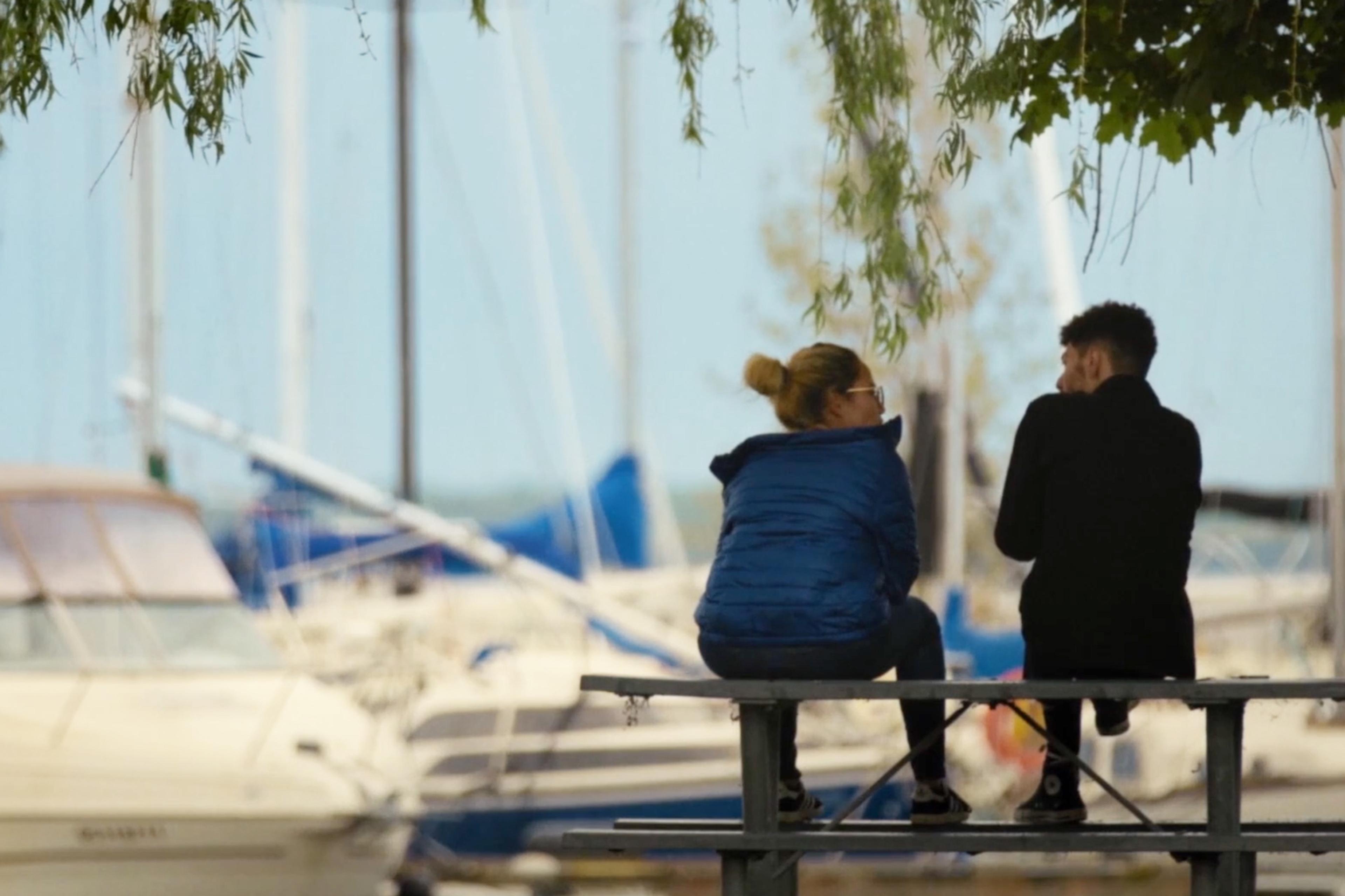 Two people sitting on a bench next to a marina with several boats in the background, partially covered by tree branches.