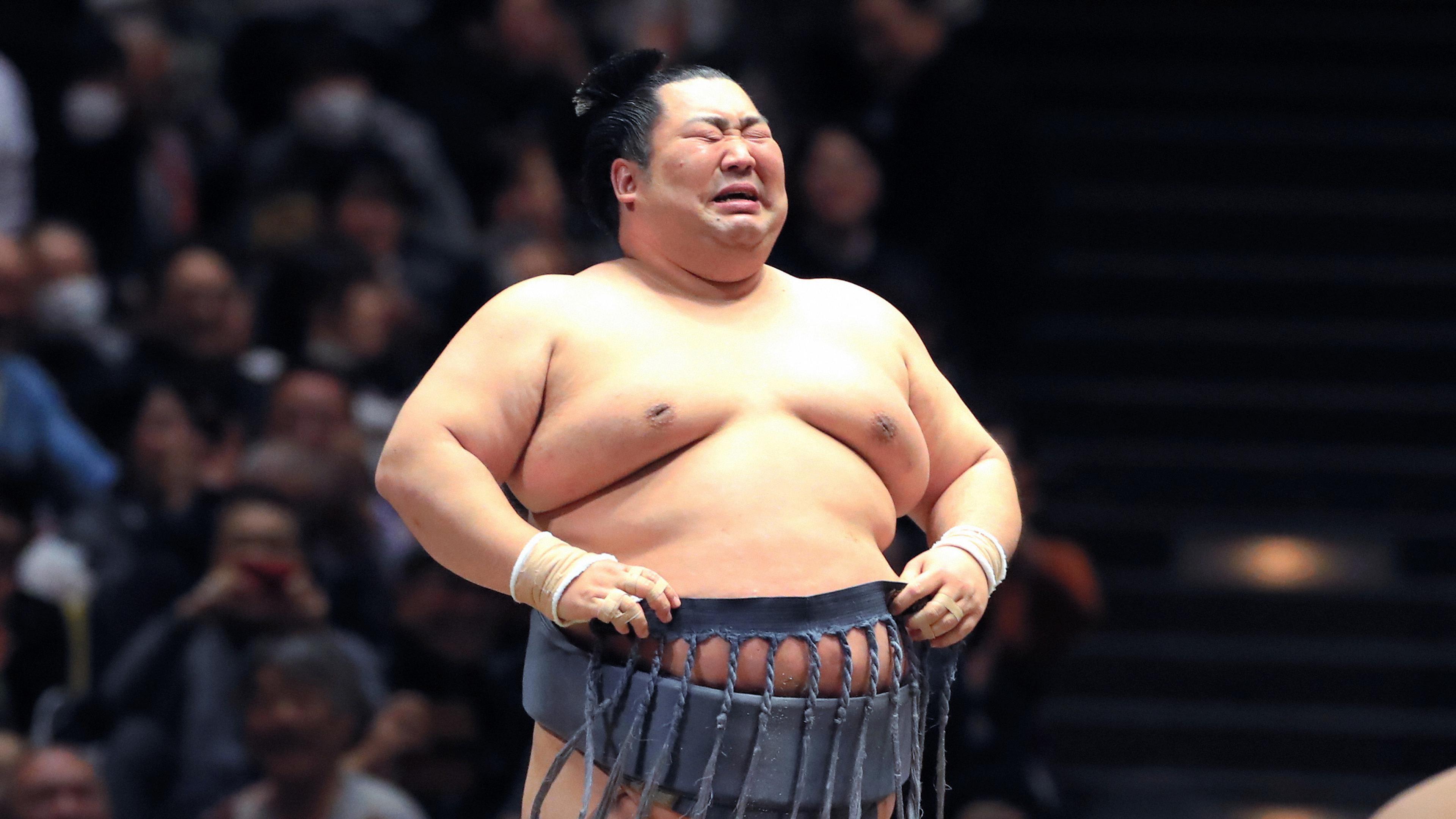 Photo of a sumo wrestler in traditional attire, making a facial expression, with an audience in the background.