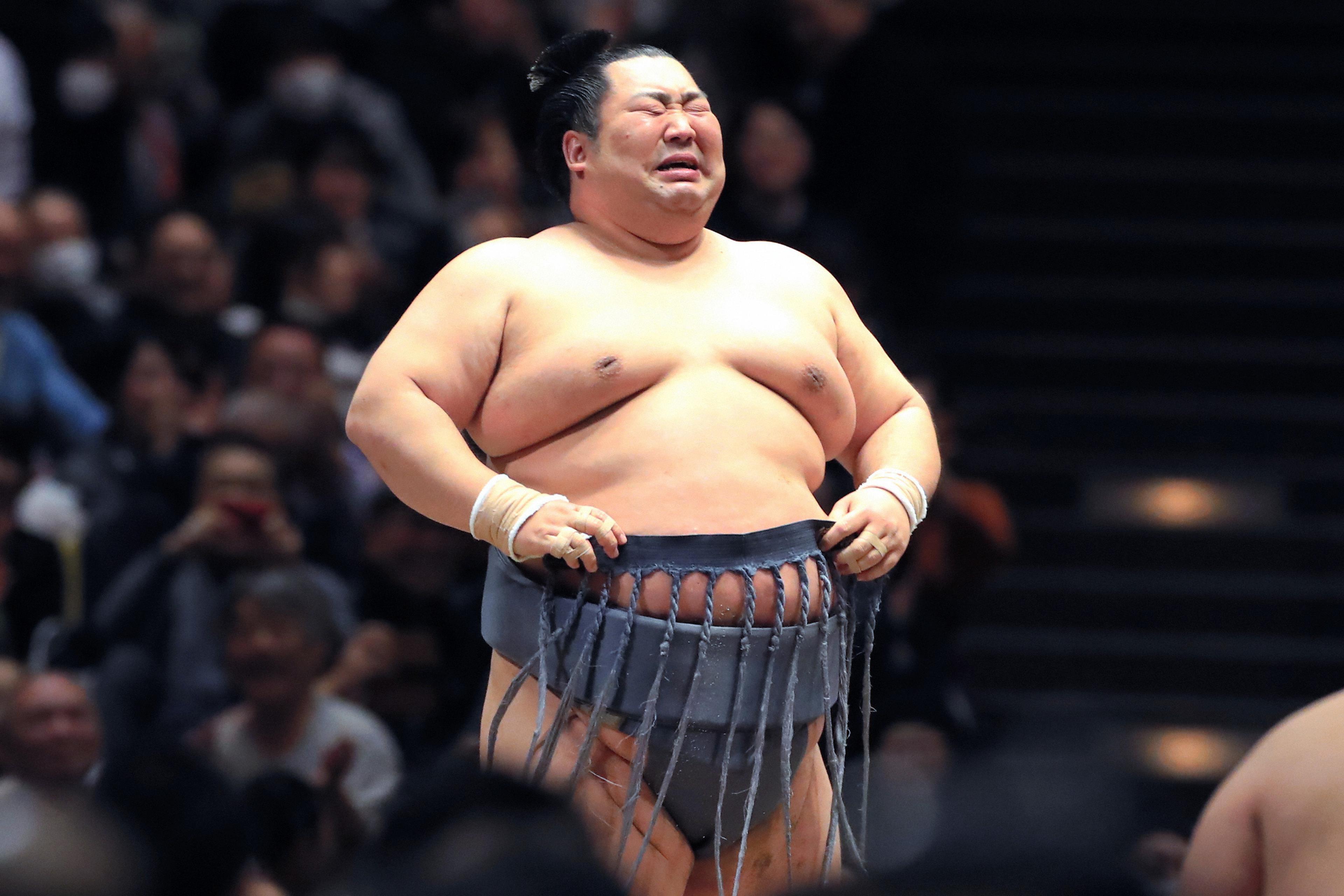 Photo of a sumo wrestler in traditional attire, making a facial expression, with an audience in the background.