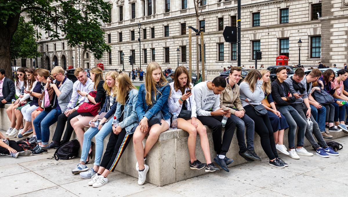 Photo of a group of young people in casual clothing sitting on a concrete bench outside a historic building, some using phones.