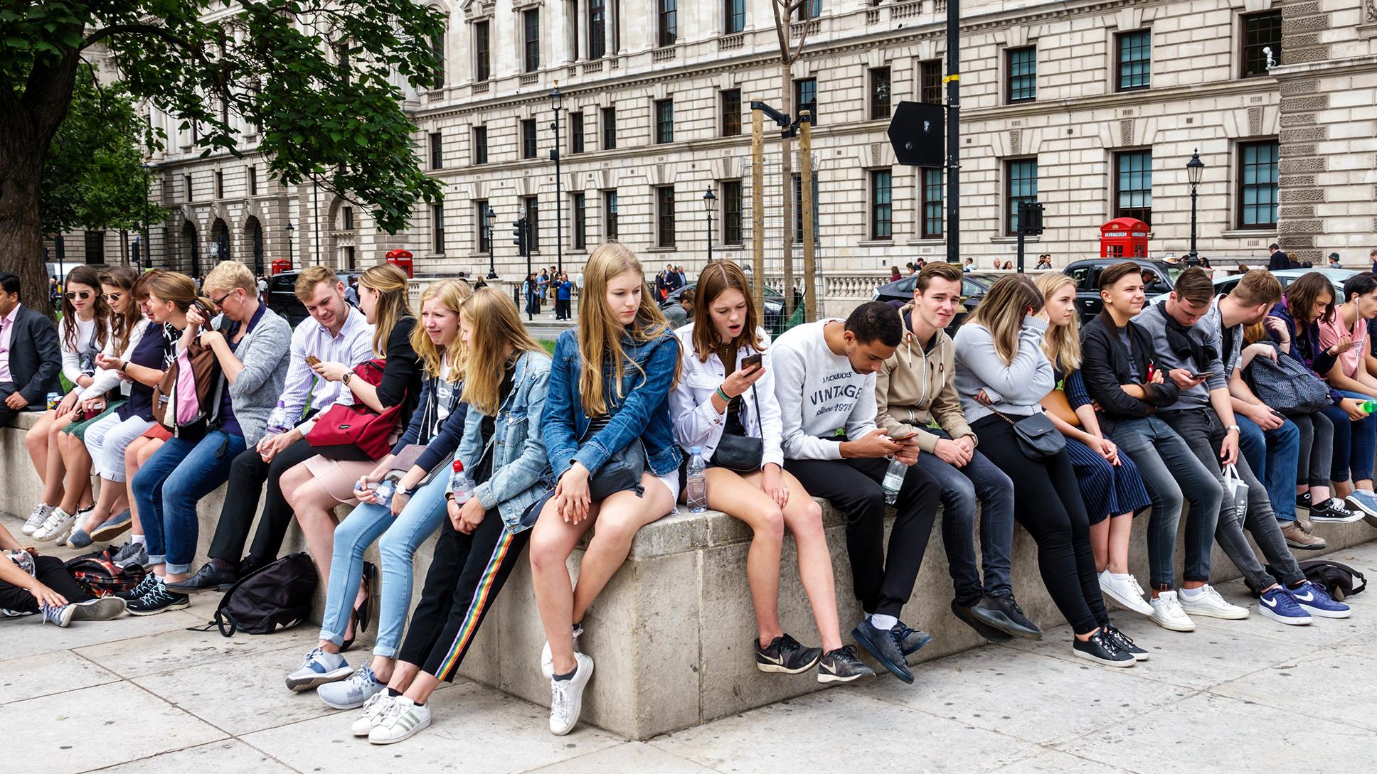 Photo of a group of young people in casual clothing sitting on a concrete bench outside a historic building, some using phones.