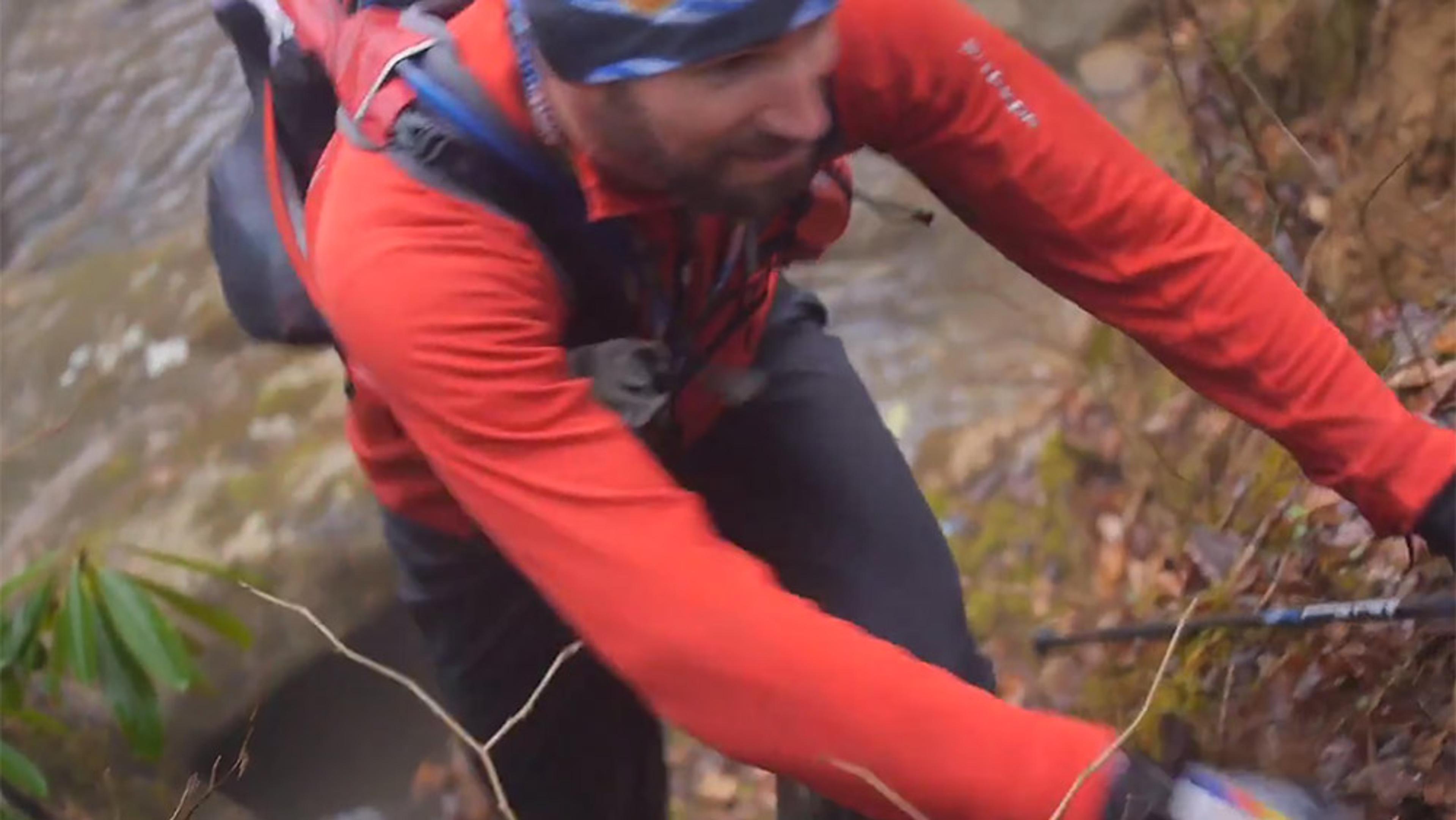 A man in outdoor gear and a red jacket scrambling up a slope in a forested area.