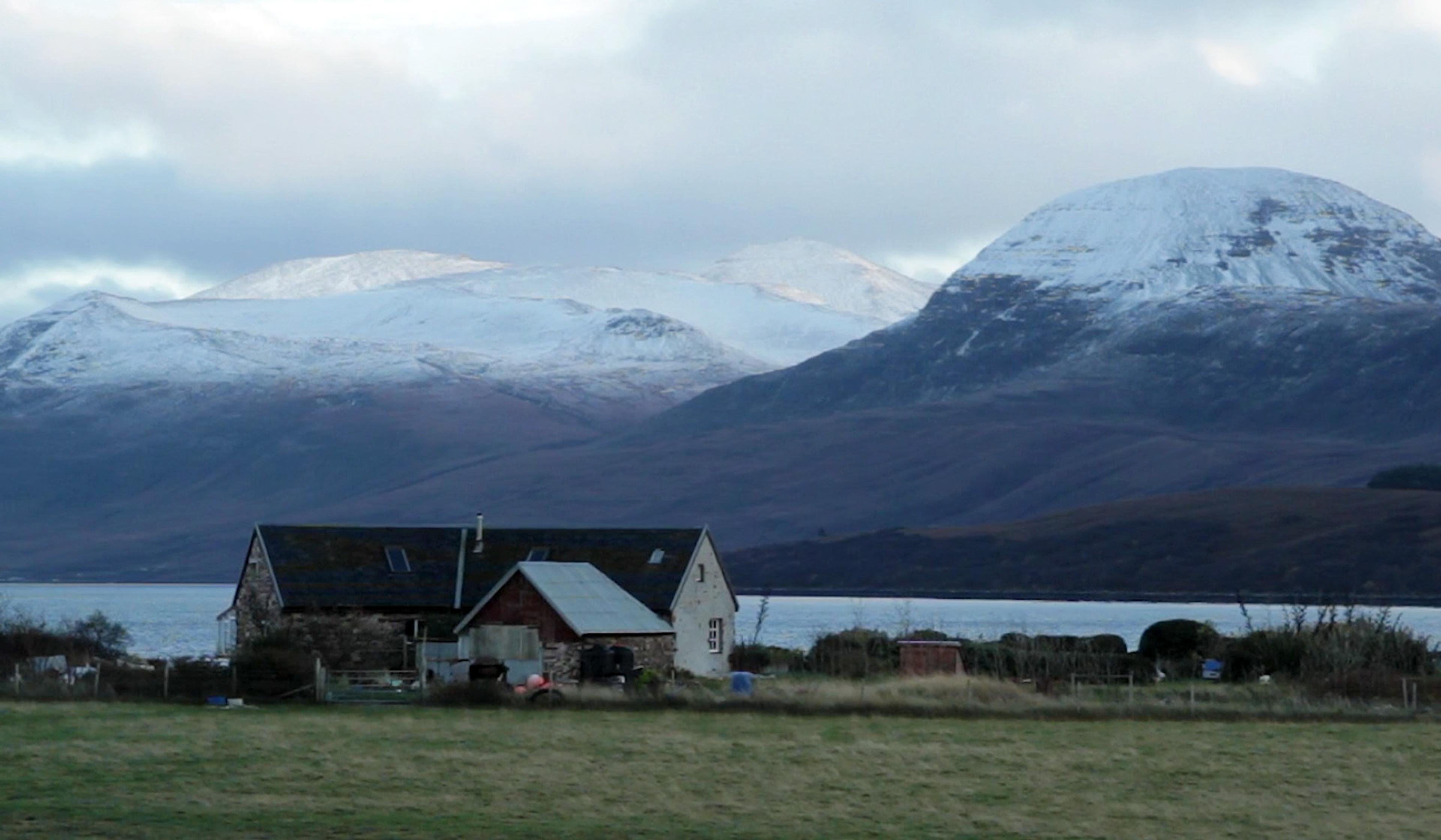 A rustic house near a lake with snow-capped mountains in the background under a cloudy sky.