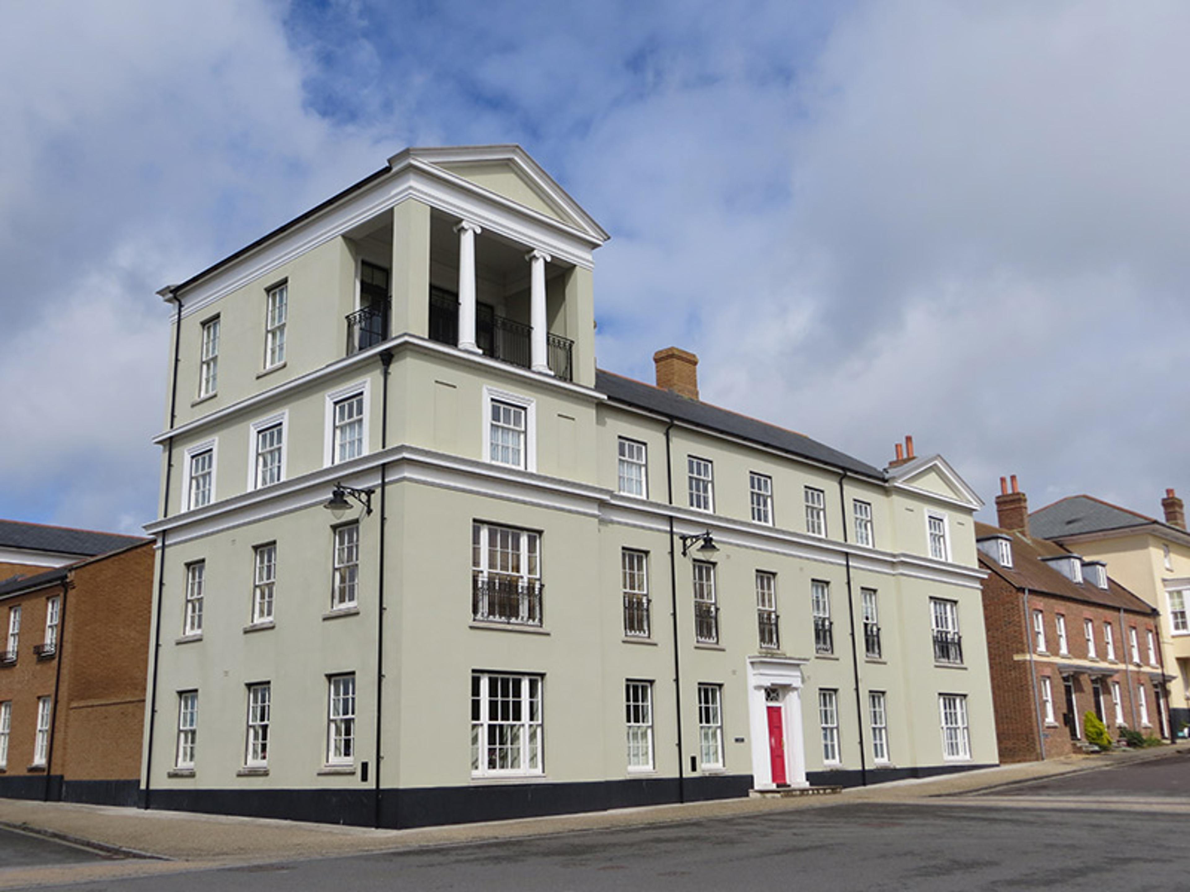 A large, light-green, three-storey building with a red door, white trim, and a columned balcony on the top floor in a residential area.
