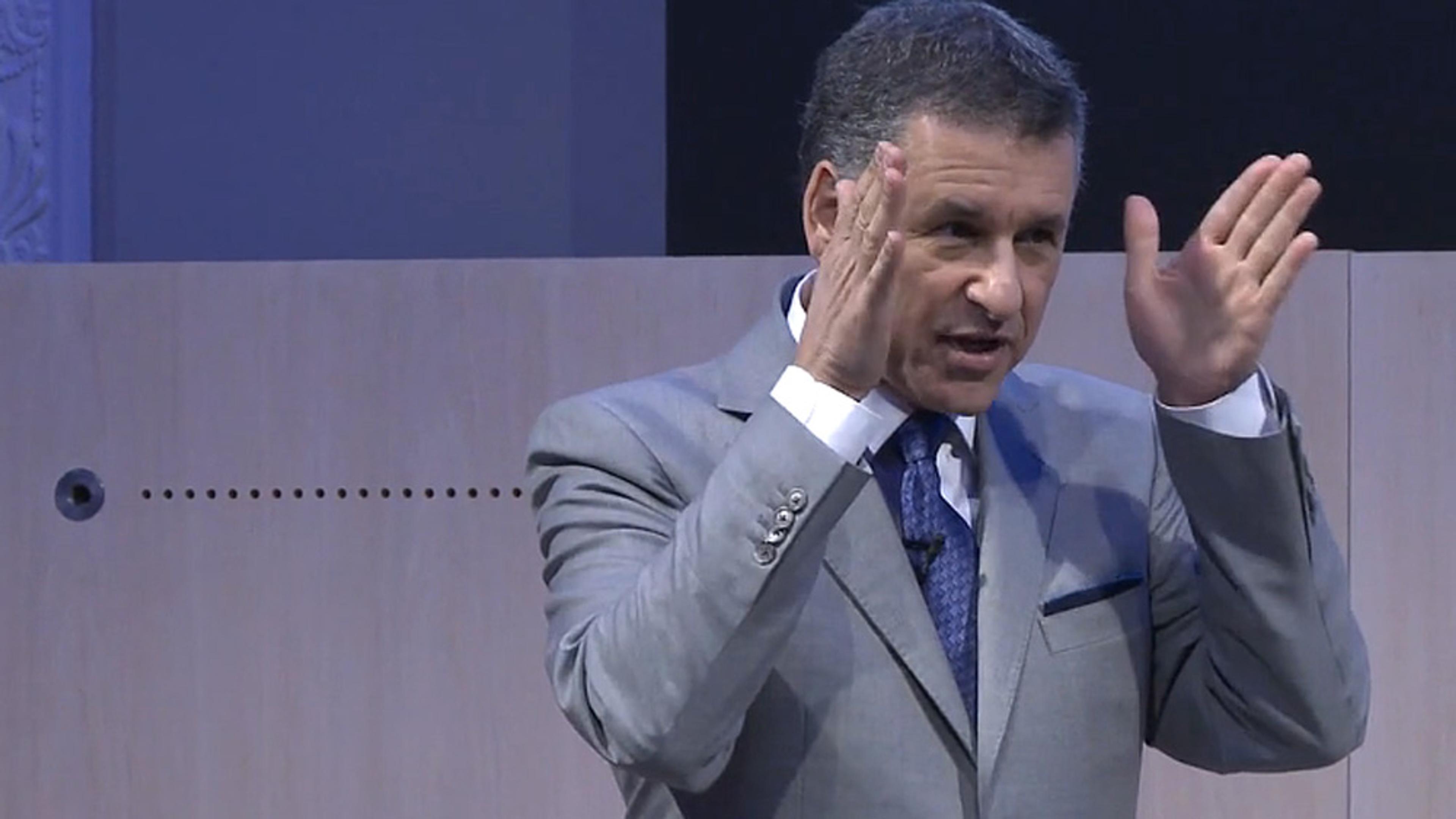 A man in a grey suit and blue tie gesturing with both hands raised, speaking at an indoor event with a wooden backdrop.