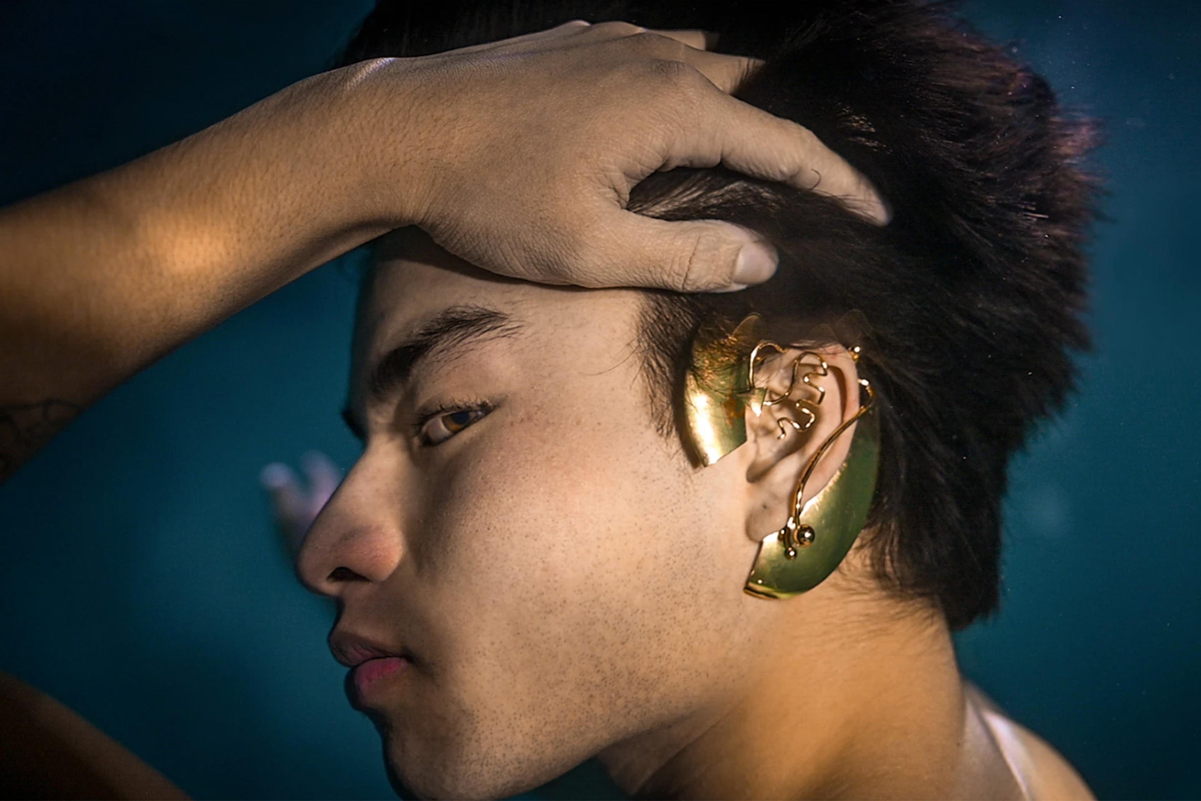 A young man turns his head with his hand to reveal a unique gold ear accessory.