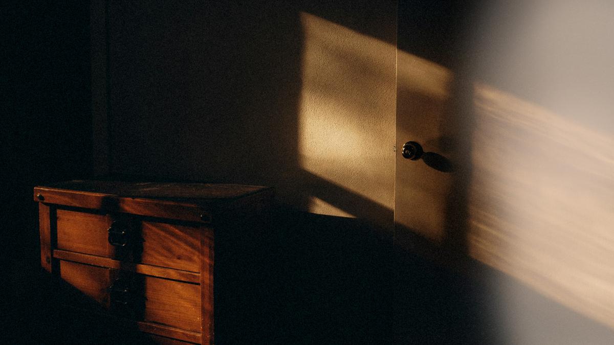 Warm sunlight casting shadows on a wooden chest and a door inside a dimly lit room.
